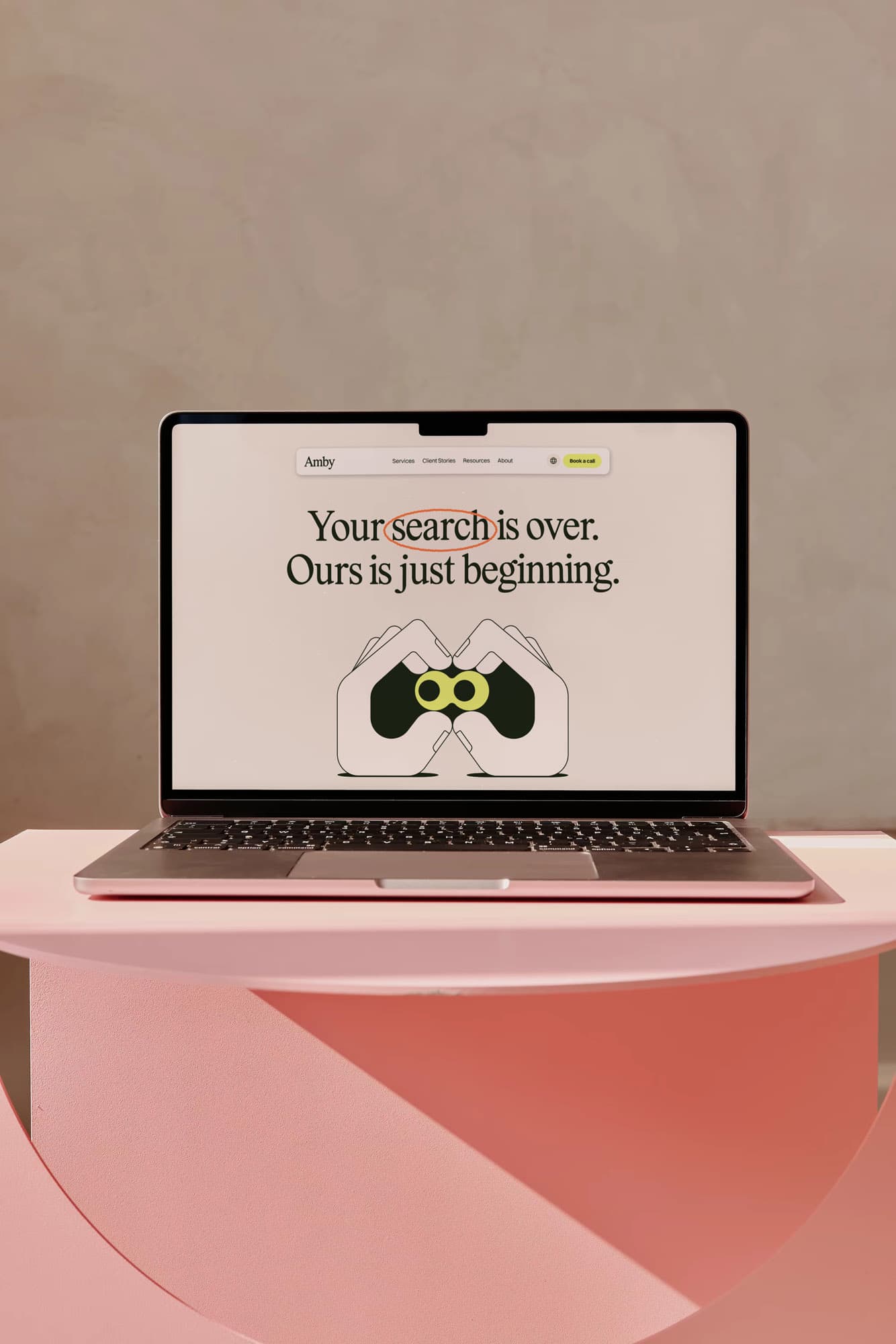 Laptop on a coral pedestal displays Amby's new website homepage with the headline "Your search is over. Ours is just beginning." and an illustrated icon of hands forming binoculars – from Duties' brand identity for recruitment agency Amby.