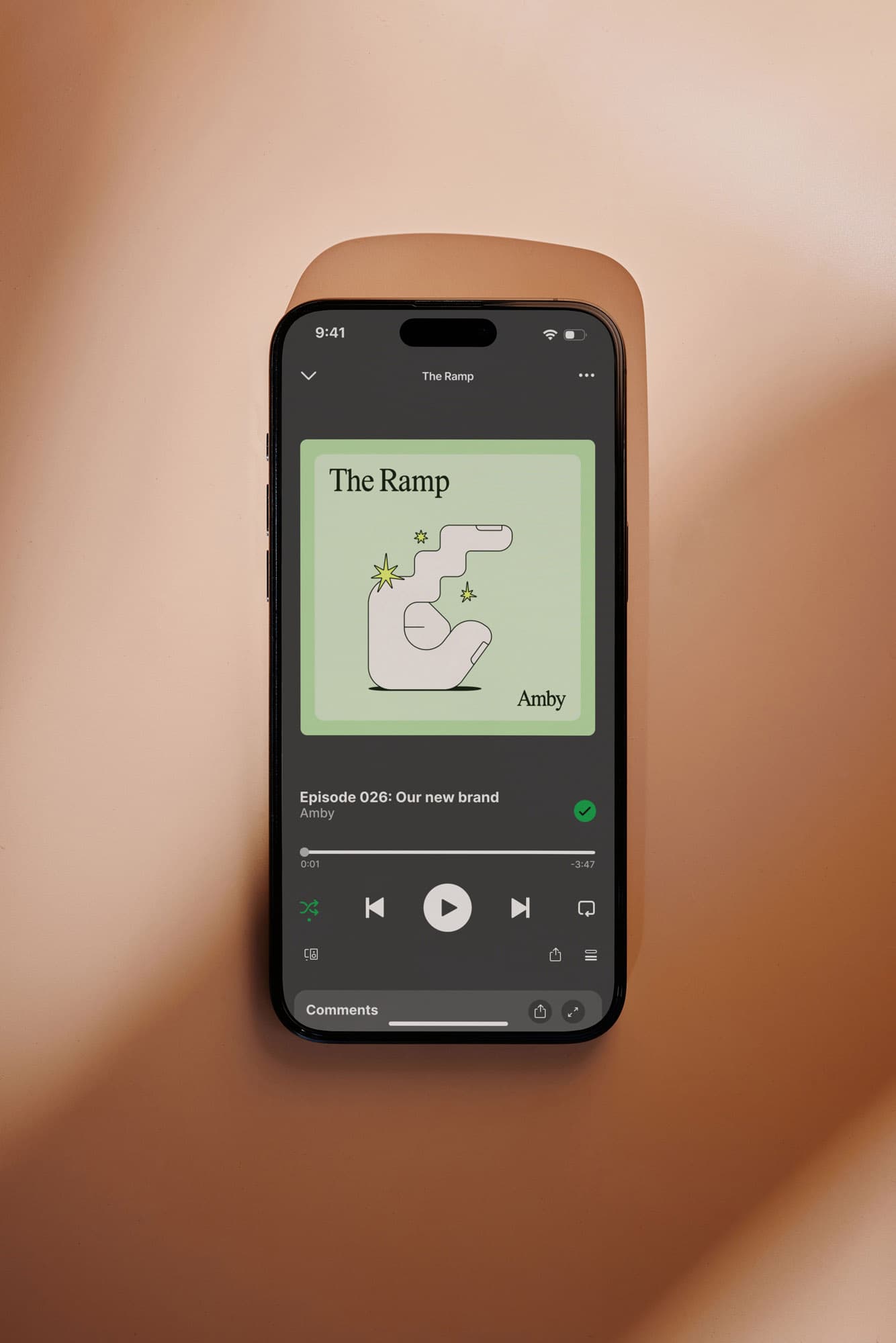 Smartphone on an orange surface displays a podcast player showing "The Ramp" cover art – an illustrated arm with stars – and the episode title "Episode 026: Our new brand" from Amby.