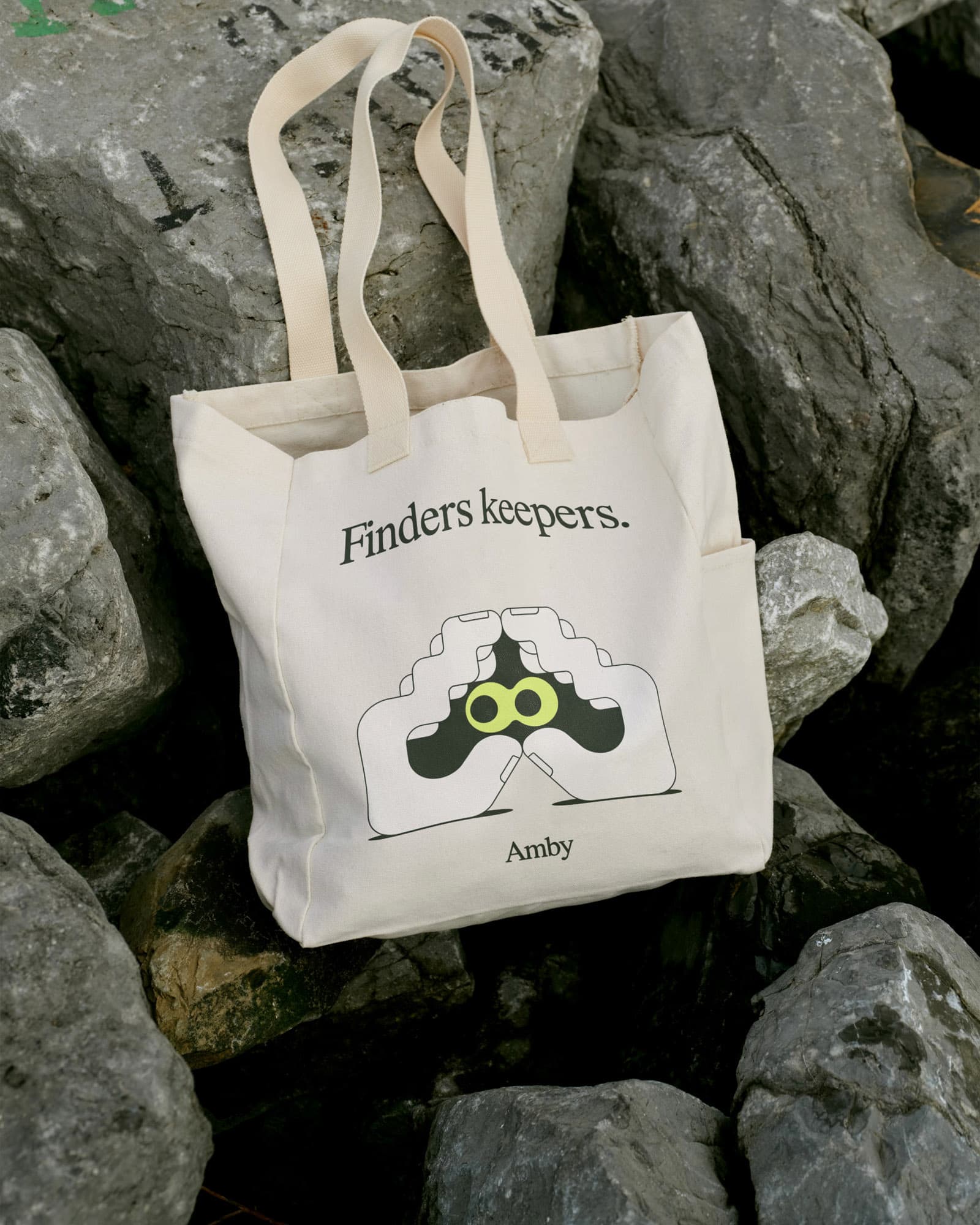 The same "Finders keepers." tote bag with the binoculars icon, photographed from a different angle on rocks.