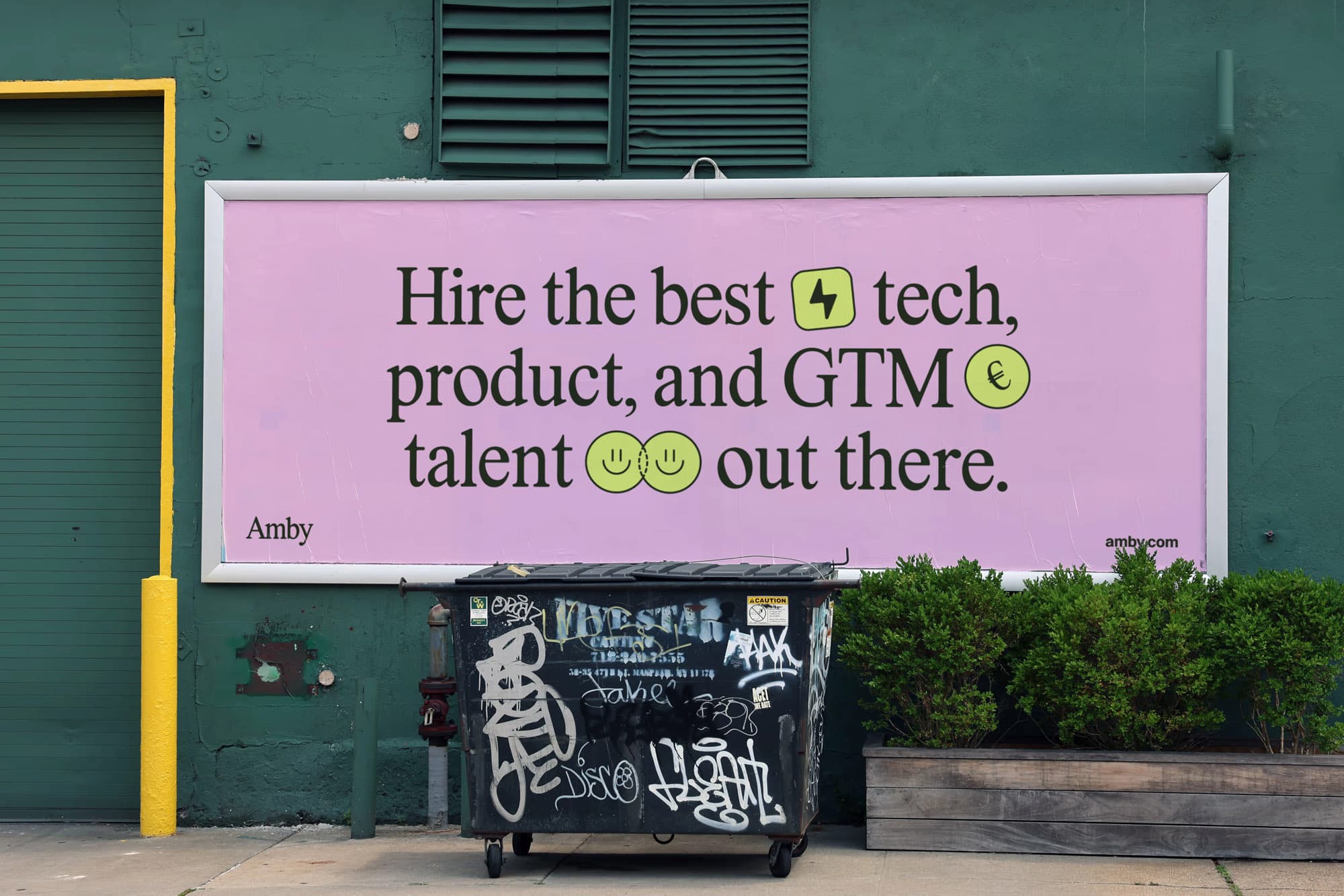 Large billboard mounted on a green building wall above a dumpster. The pink ad reads "Hire the best [lightning bolt icon] tech, product, and GTM [coin icon] talent [smiley icons] out there." with the Amby logo and amby.com.