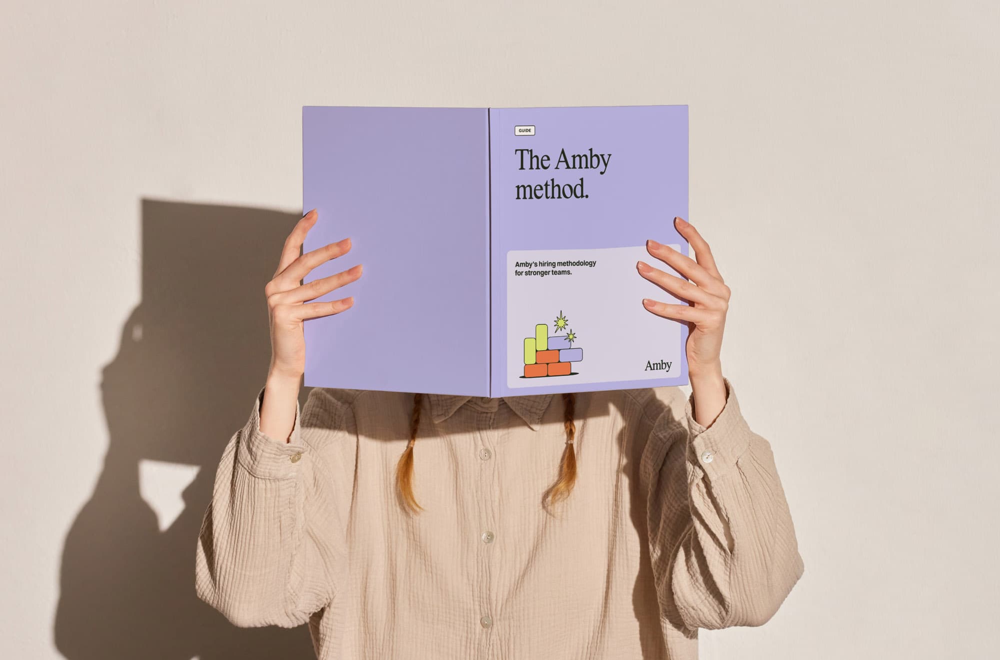 A person holds up an open lavender guide titled "The Amby method." with the subtitle "Amby's hiring methodology for stronger teams.", featuring an illustration of bricks and a diamond, and the Amby logo.