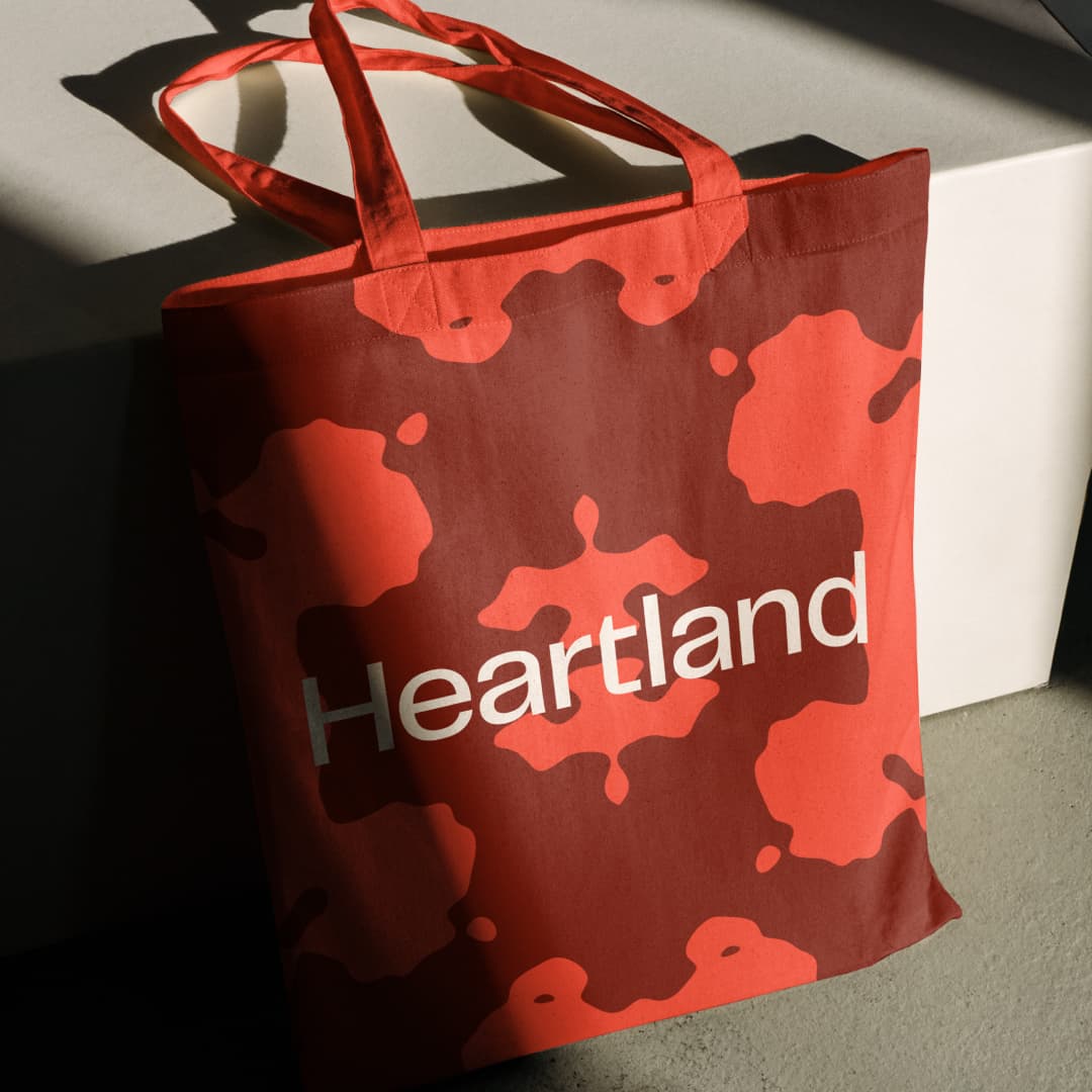 Red Heartland tote bag with paper clip-inspired pattern in two shades behind white wordmark in dramatic directional sunlight.