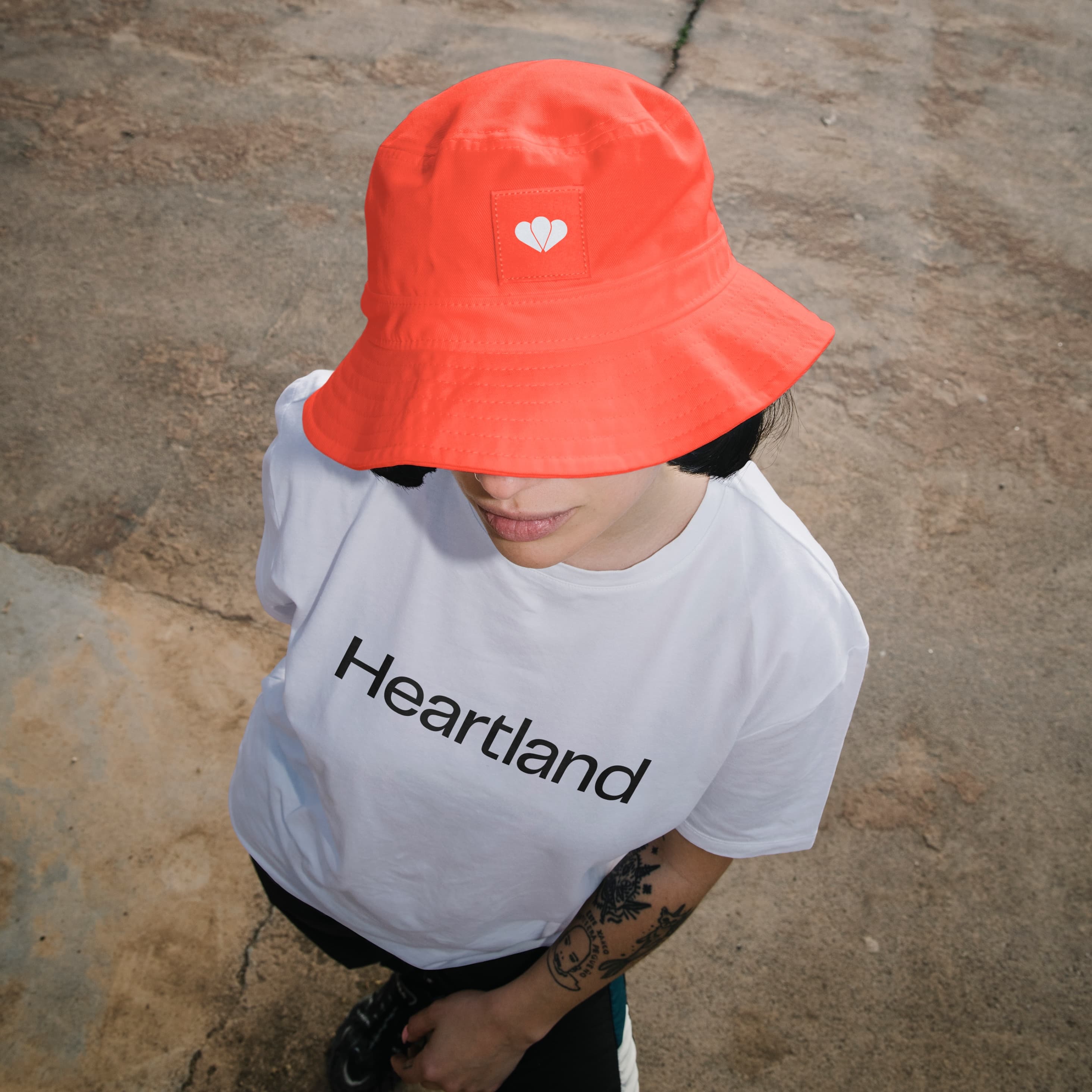 Merchandise photo shot from above: person wearing an orange Heartland bucket hat with the double-heart brandmark on a patch, and a white Heartland t-shirt.