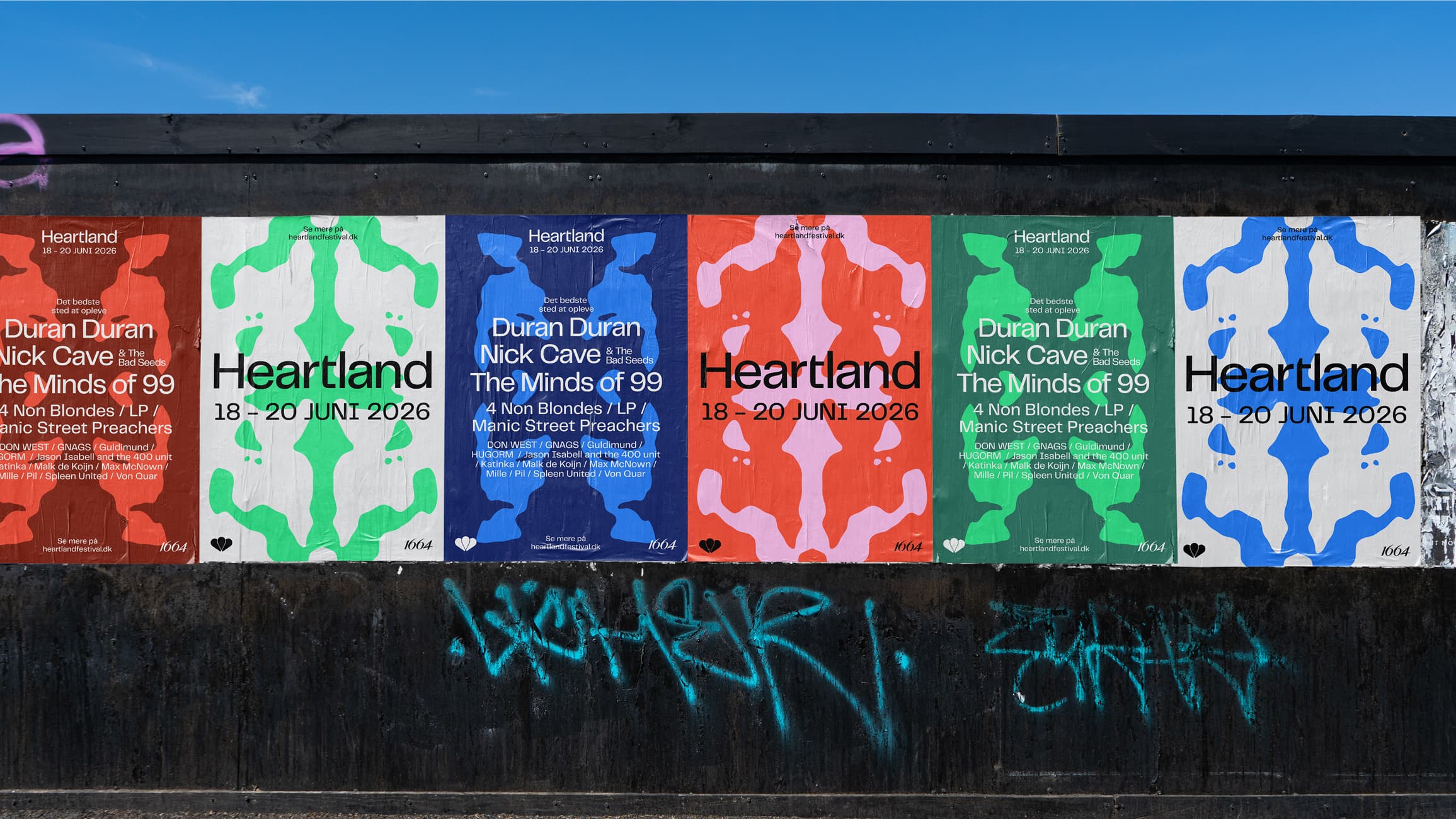 Street photography: six Heartland festival posters wheat-pasted side by side on a black wall with graffiti, in alternating colours (red, white, blue, orange, green), all showing the lineup with organic blob pattern. Festival 18–20 June 2026.