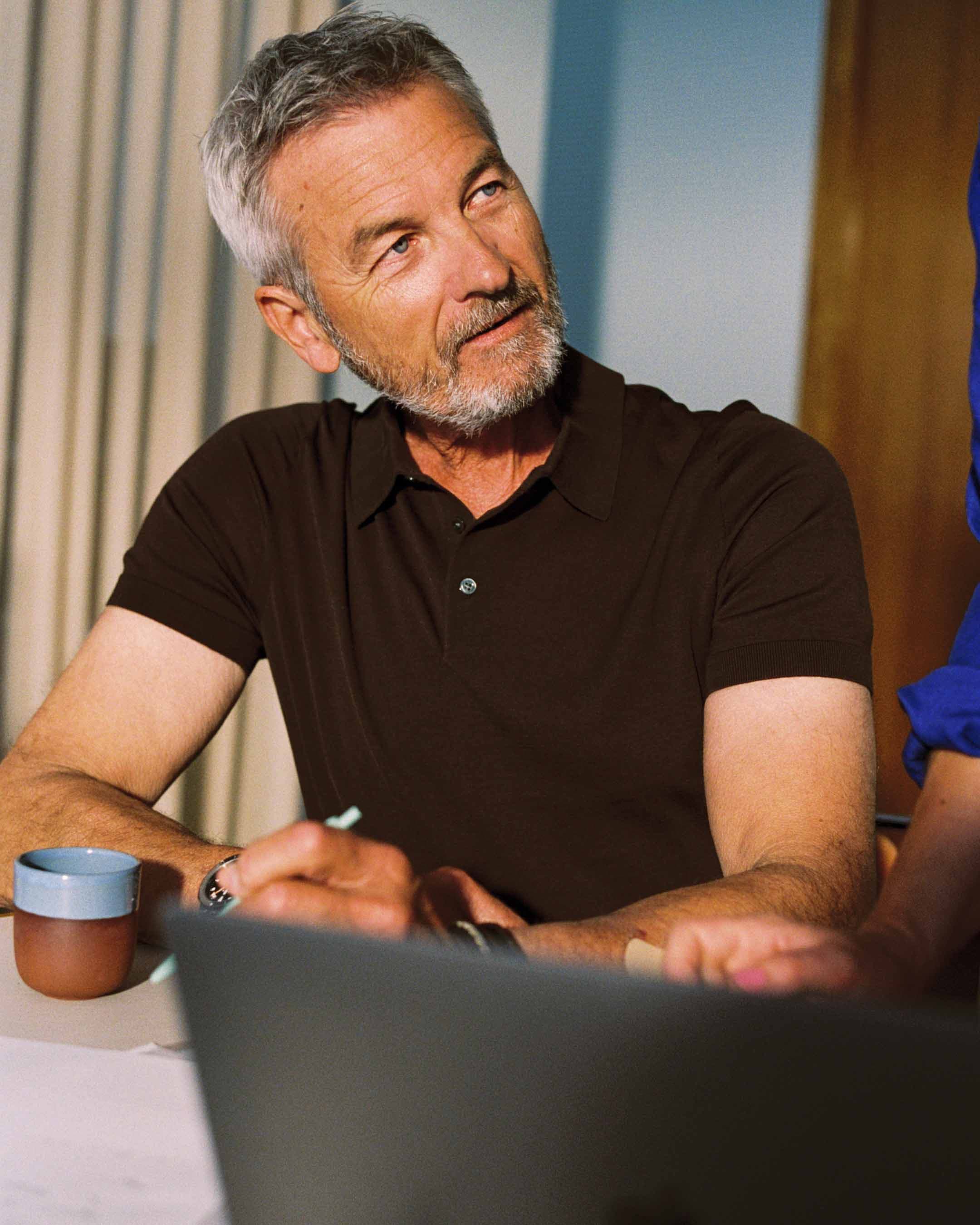 Grey-haired bearded man in a dark brown polo shirt looks thoughtfully up and to the side, seated at a table with a laptop and ceramic cup. Warm, film-like photography in NKI's new image style.