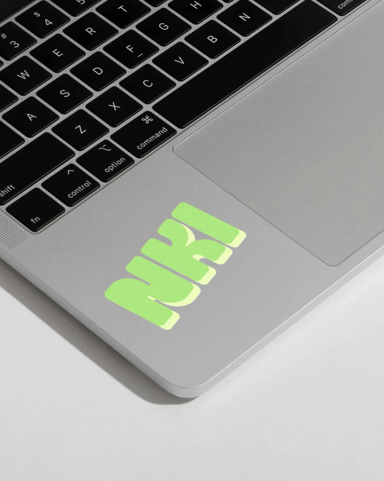 The NKI logo in rounded light-green letters with a soft shadow, applied as a sticker on the trackpad of a silver laptop. Mockup showing the brand on everyday study equipment.