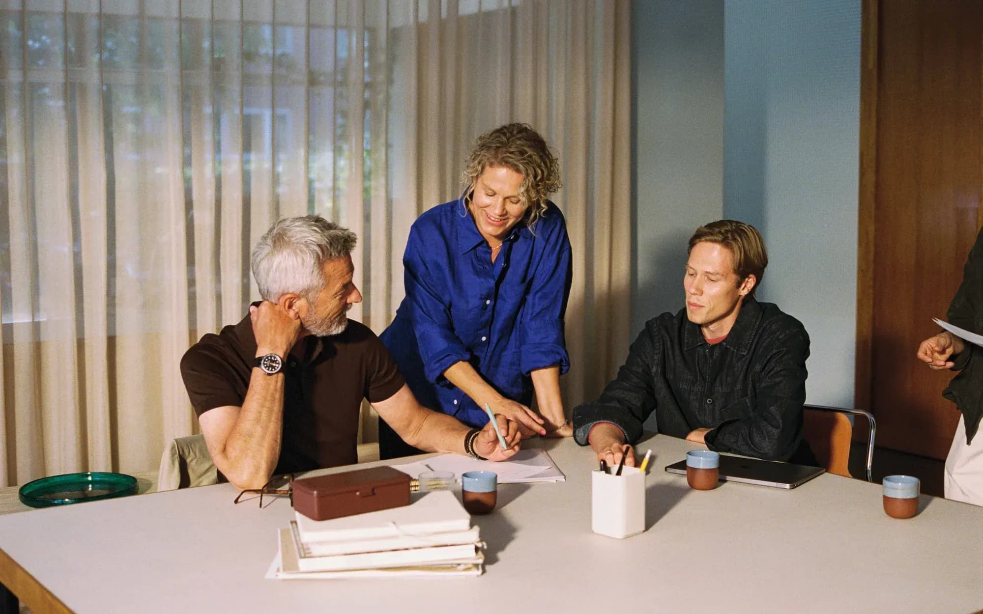Three colleagues work together at a white table: a grey-haired man in a brown polo, a woman in a cobalt-blue shirt pointing at papers, and a younger man in a dark jacket. Calm, film-style office scene.