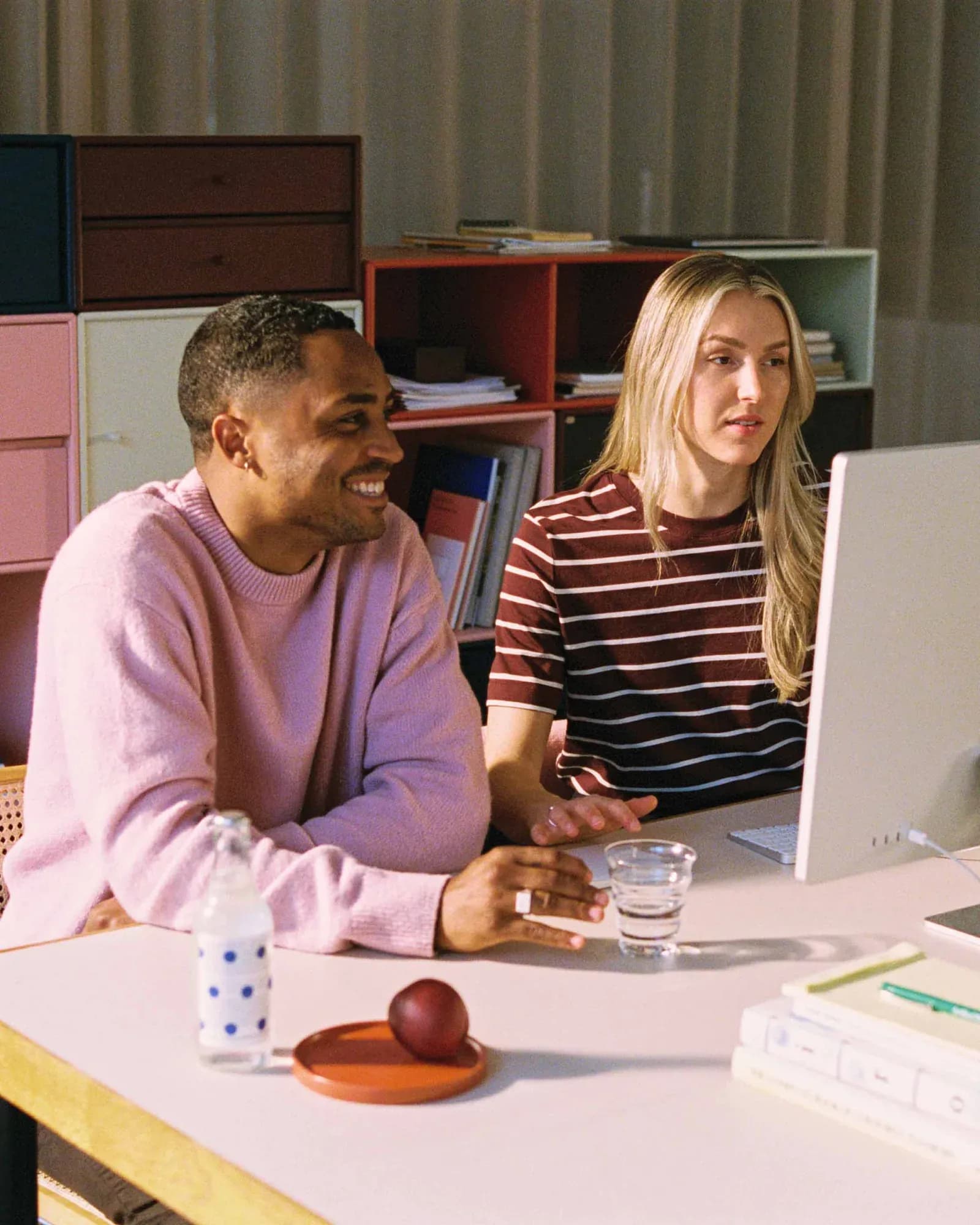 A smiling man in a pink knit sweater and a woman in a burgundy striped t-shirt look at a monitor at a desk. Colourful storage modules fill the background; a bottle and a piece of fruit sit on the desk.