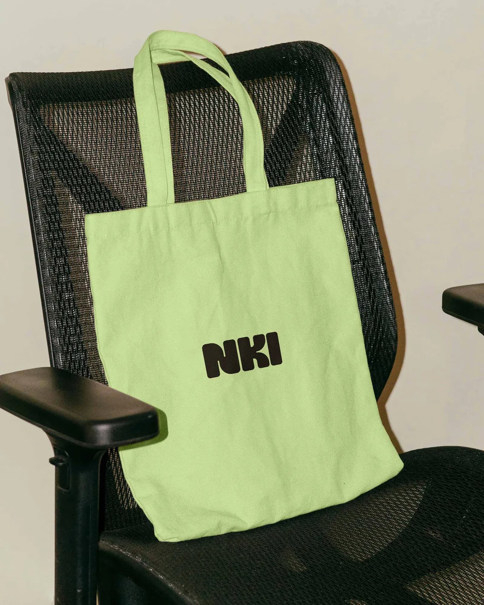 Light-green canvas tote bag with the black NKI logo hanging over the back of a black mesh office chair. Branded merchandise in the brand's signature green.