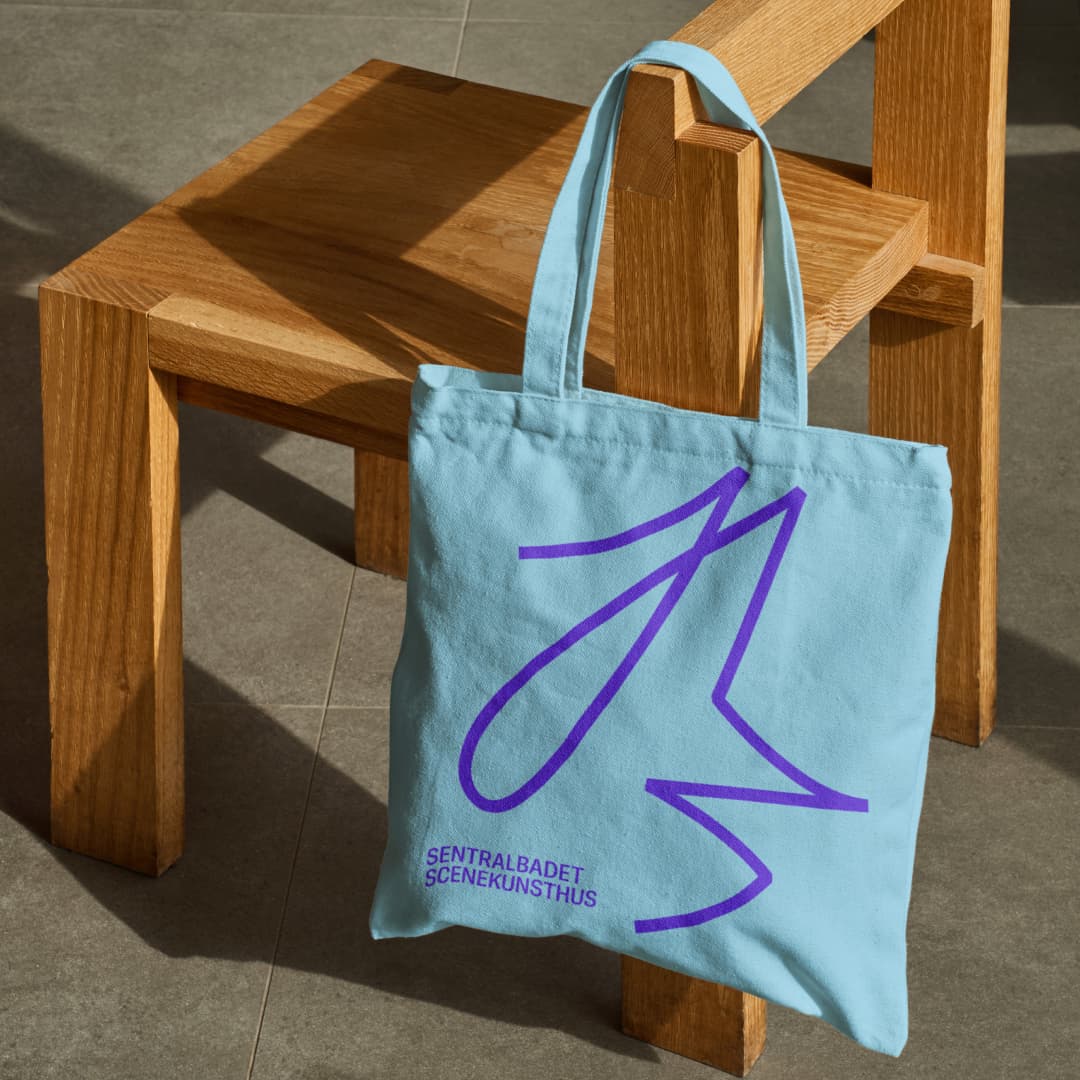 Light blue tote bag featuring Sentralbadet Scenekunsthus' purple logo and wordmark, hanging on a wooden chair – an example of merchandise within the new visual identity.