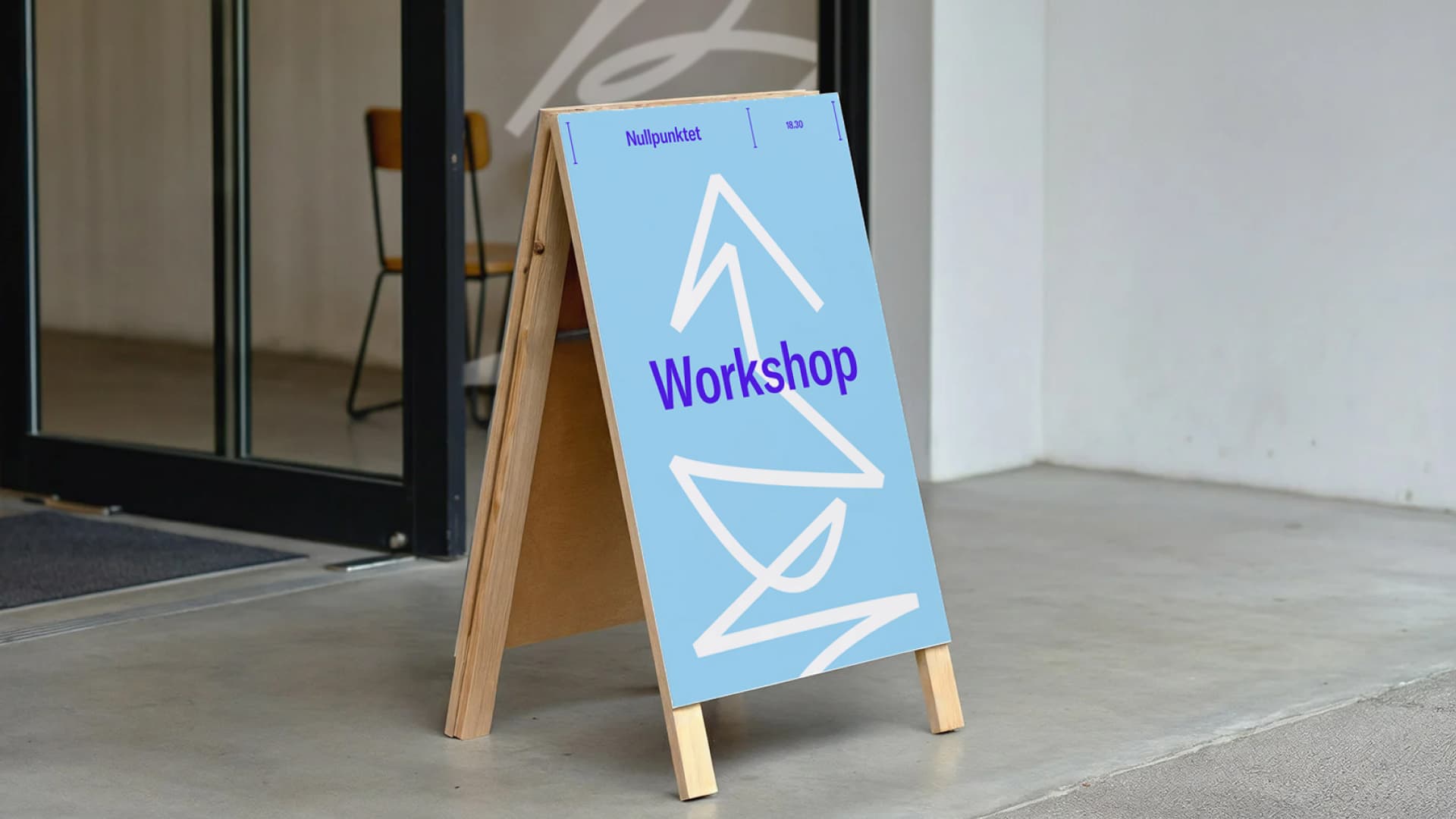 A-frame sandwich board outside an entrance displaying a light blue poster for a 'Workshop' at 18:30 ('Nullpunktet'), decorated with white line drawings from Sentralbadet Scenekunsthus' visual identity.