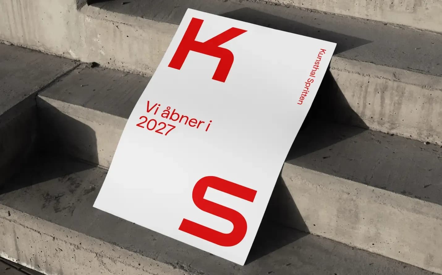 White poster with red KS logo and the text 'Vi åbner i 2027' leaning against concrete steps – opening announcement mockup.