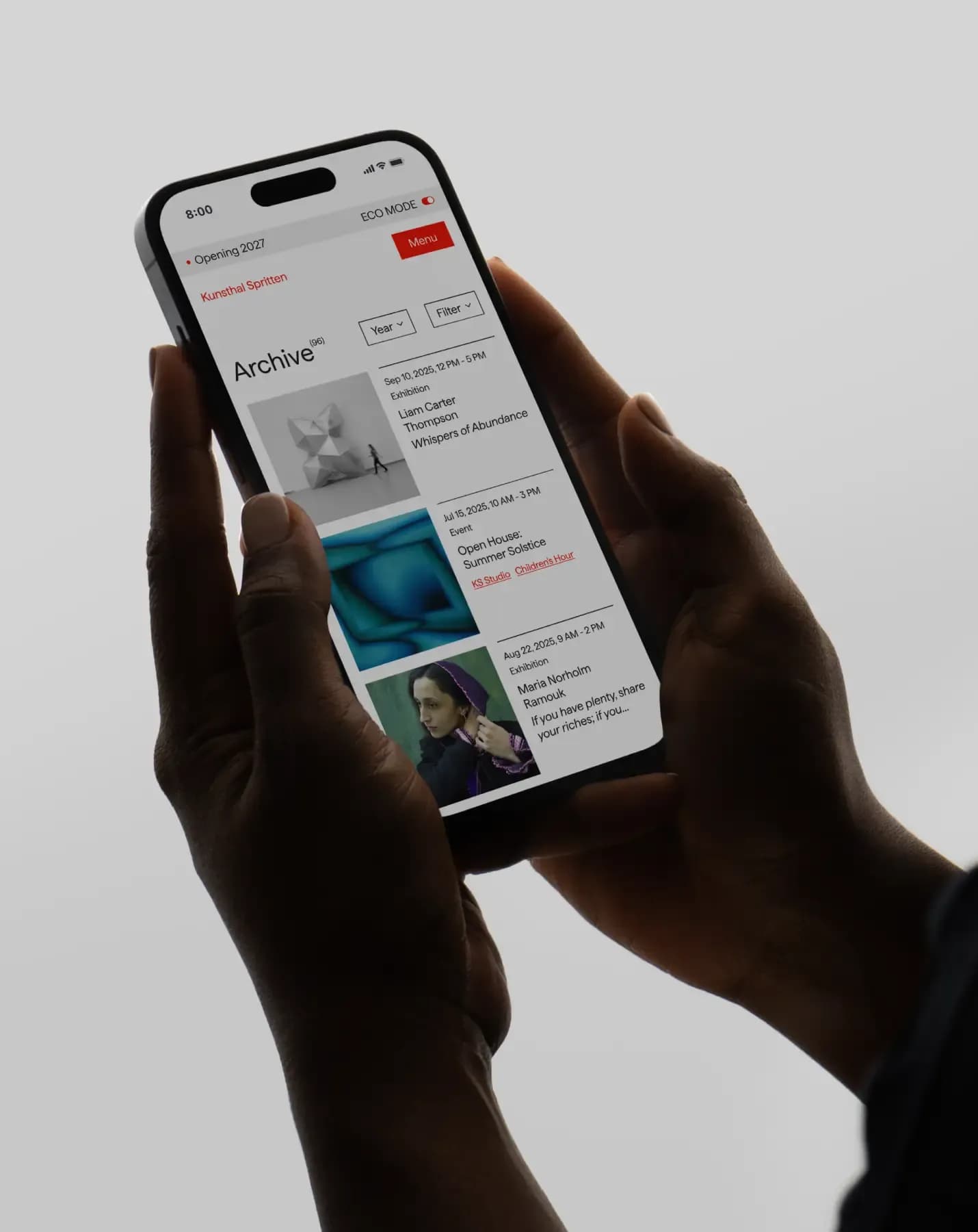 Hands holding an iPhone displaying Kunsthal Spritten's archive page with exhibition and event history and an eco mode toggle.