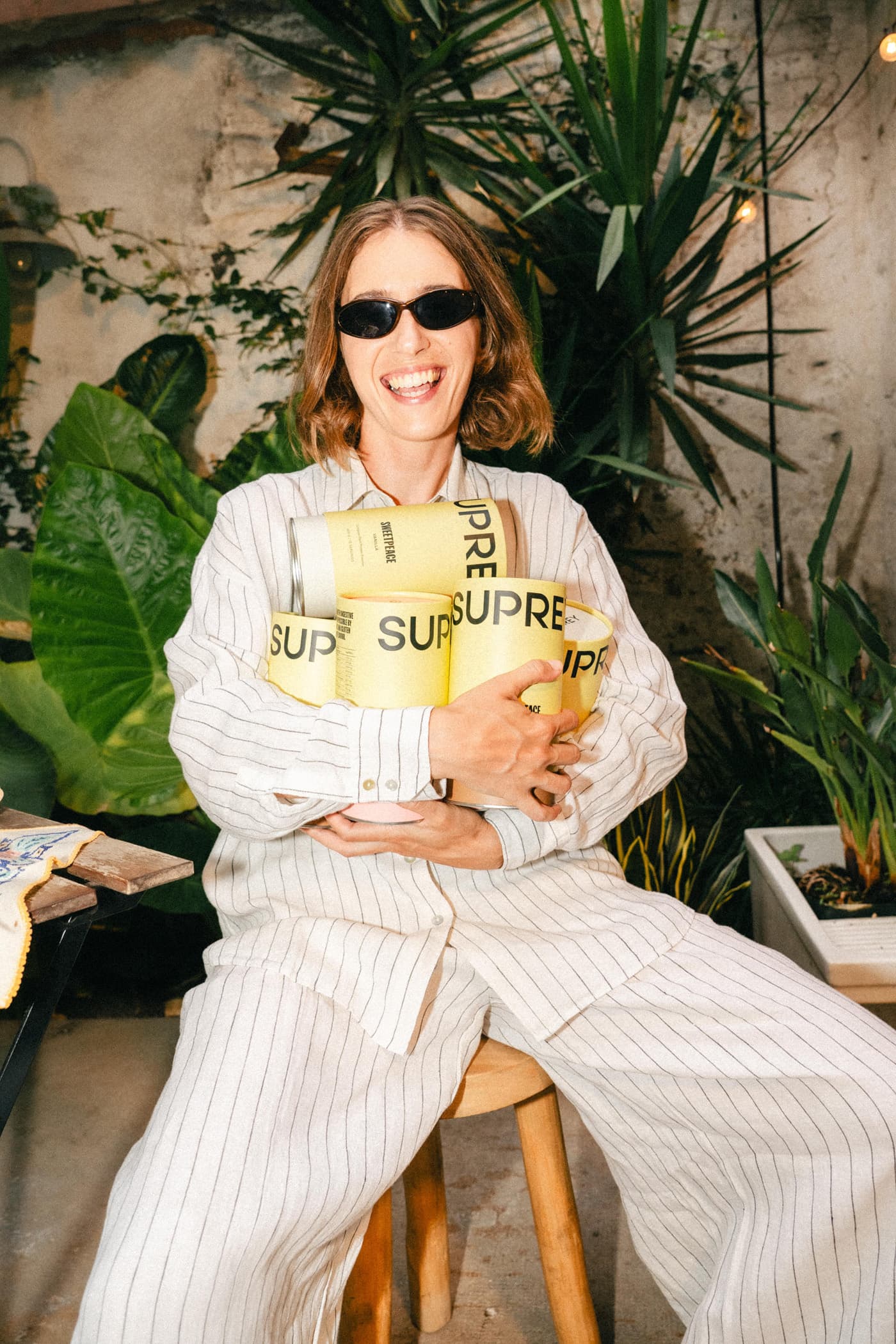 Smiling woman in pinstripe suit and sunglasses holds several yellow Suprey canisters in her arms, surrounded by green plants.