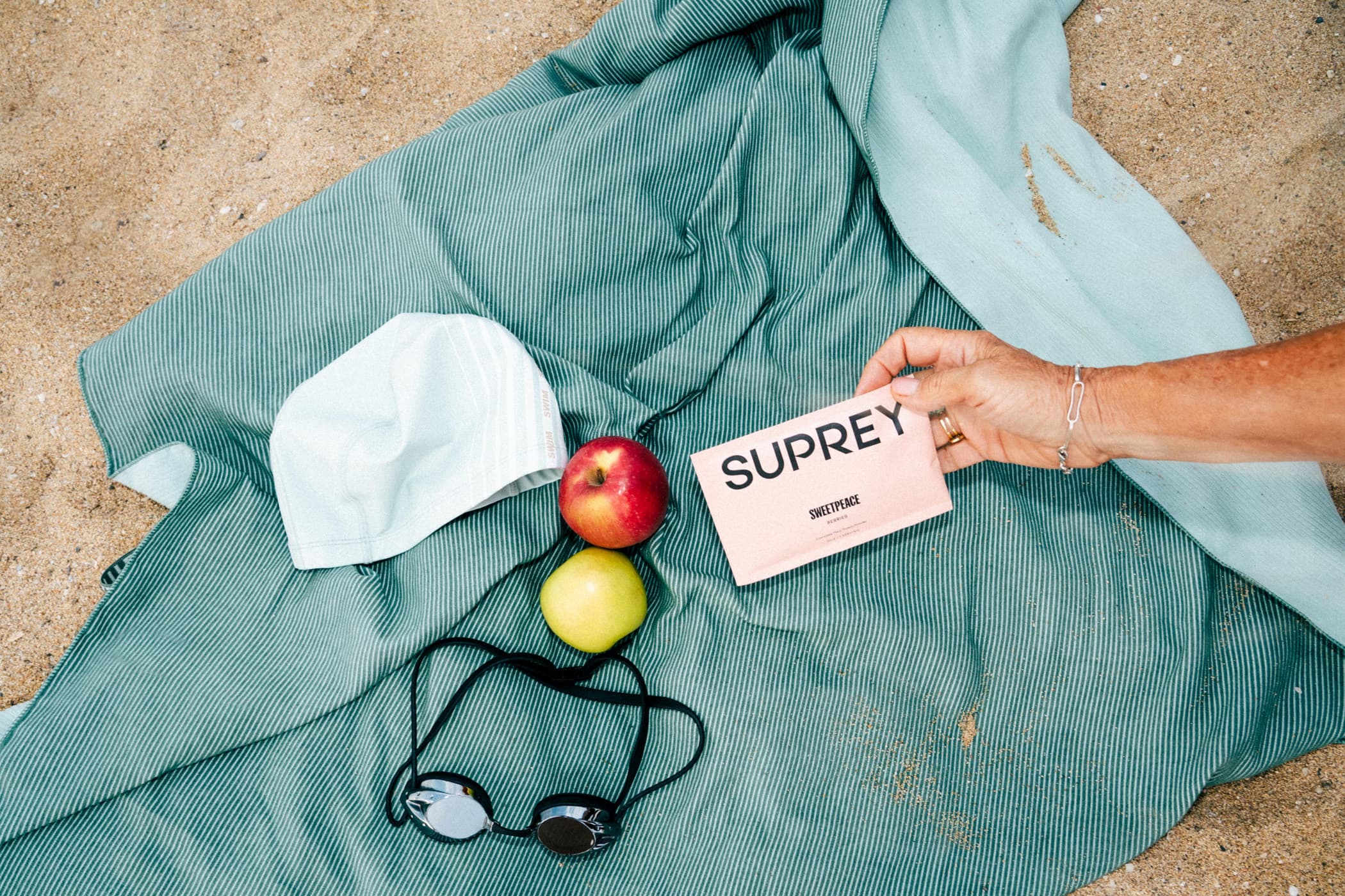 Flat lay on beach towel with swim gear: swim cap, goggles, two apples and a Suprey Sweetpeace protein packet on sand.