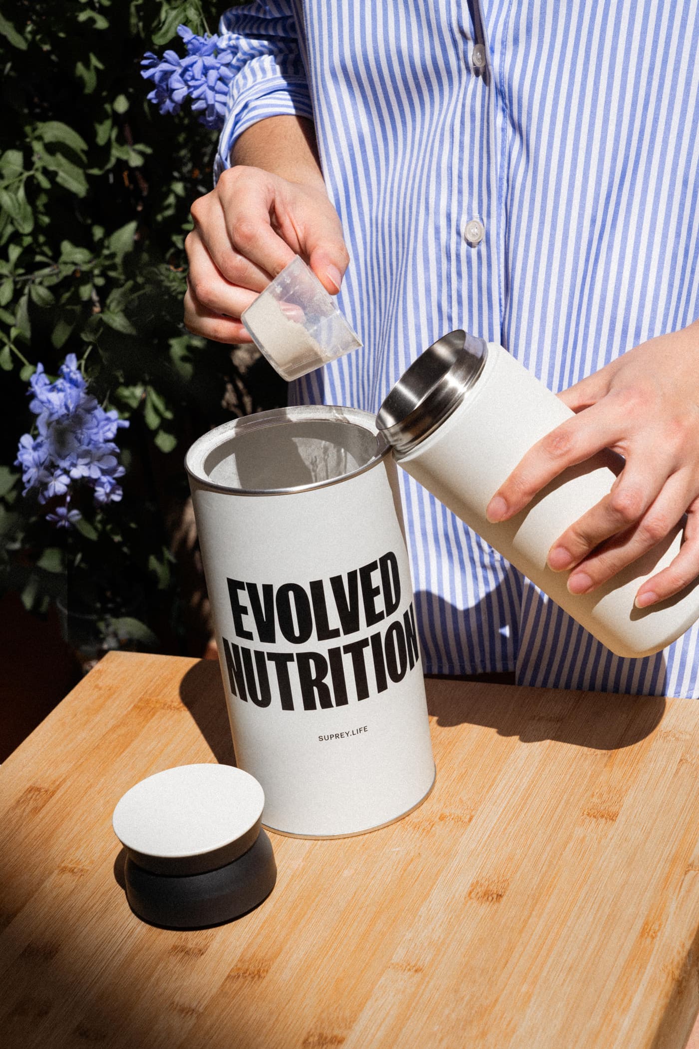 Person scoops protein powder from a Suprey Life Evolved Nutrition canister into a travel cup in sunlight. Blue-striped shirt.