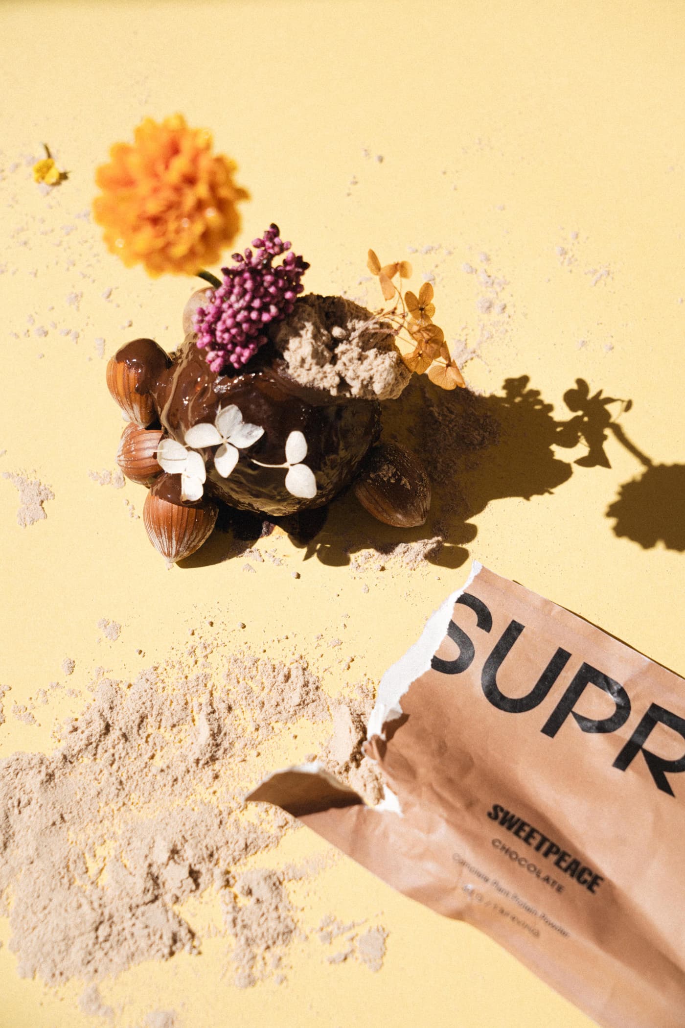 Overhead still life: chocolate, hazelnuts, flowers and scattered protein powder with torn Suprey Sweetpeace Chocolate packet on yellow background.