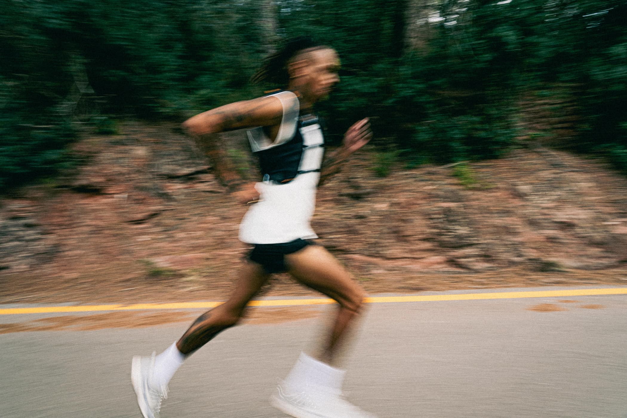 Motion-blurred runner in white top and running vest sprints on an asphalt road surrounded by dense vegetation. The image captures speed and intensity.