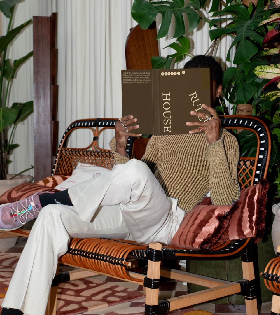 A person reclining in a rattan lounge chair among houseplants, holding up the 'House Rules' book to hide their face.