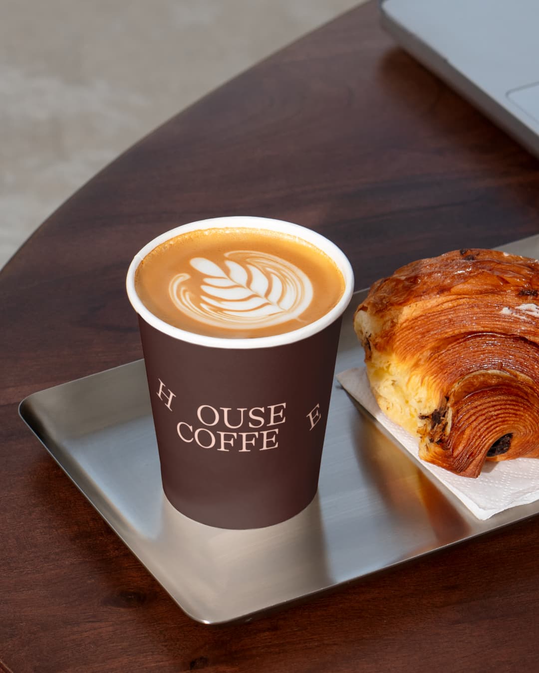 Takeaway coffee cup printed 'House Coffee' with latte art, served beside a flaky pastry on a stainless steel tray.