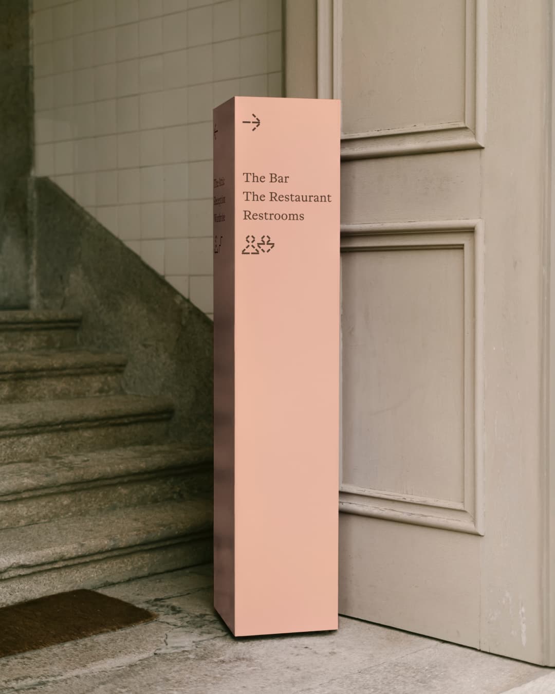Pink freestanding signage column reading 'The Bar / The Restaurant / Restrooms' with directional icons, placed beside a stone staircase.