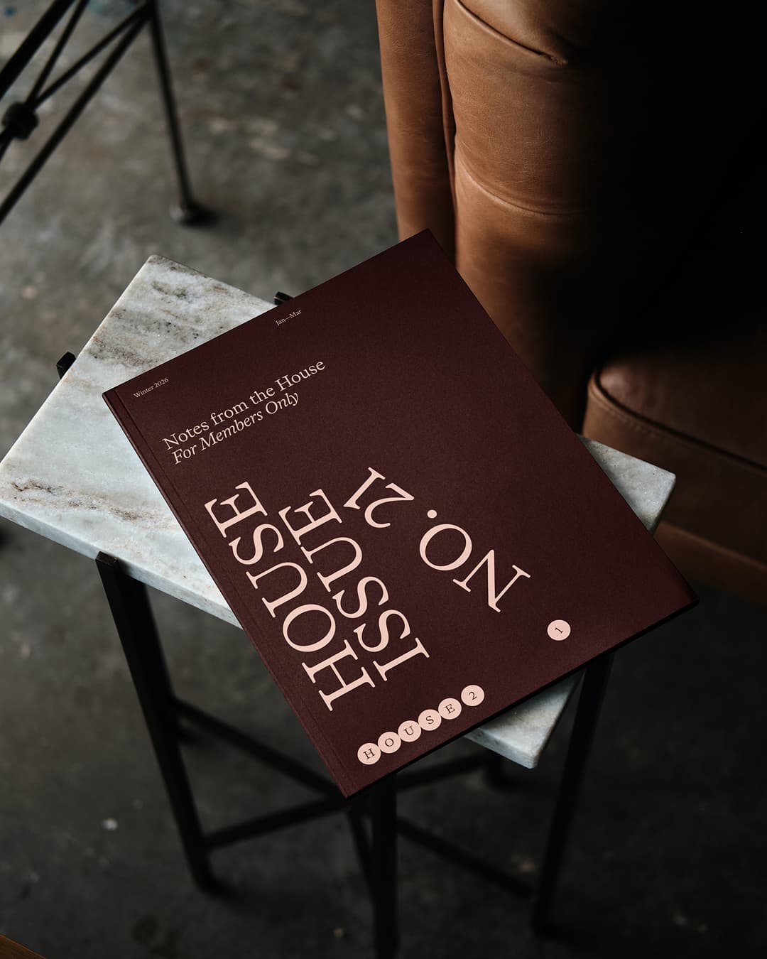 Vertical-format shot of the 'House Issue No.21' members' magazine in dark maroon, resting on a white marble side table near a leather armchair.