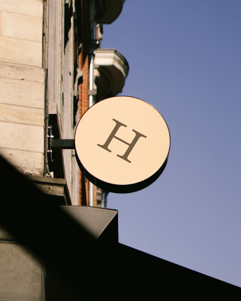Circular cream 'H' signage disc mounted on a stone building corner against a clear blue sky, House21's freestanding street sign.