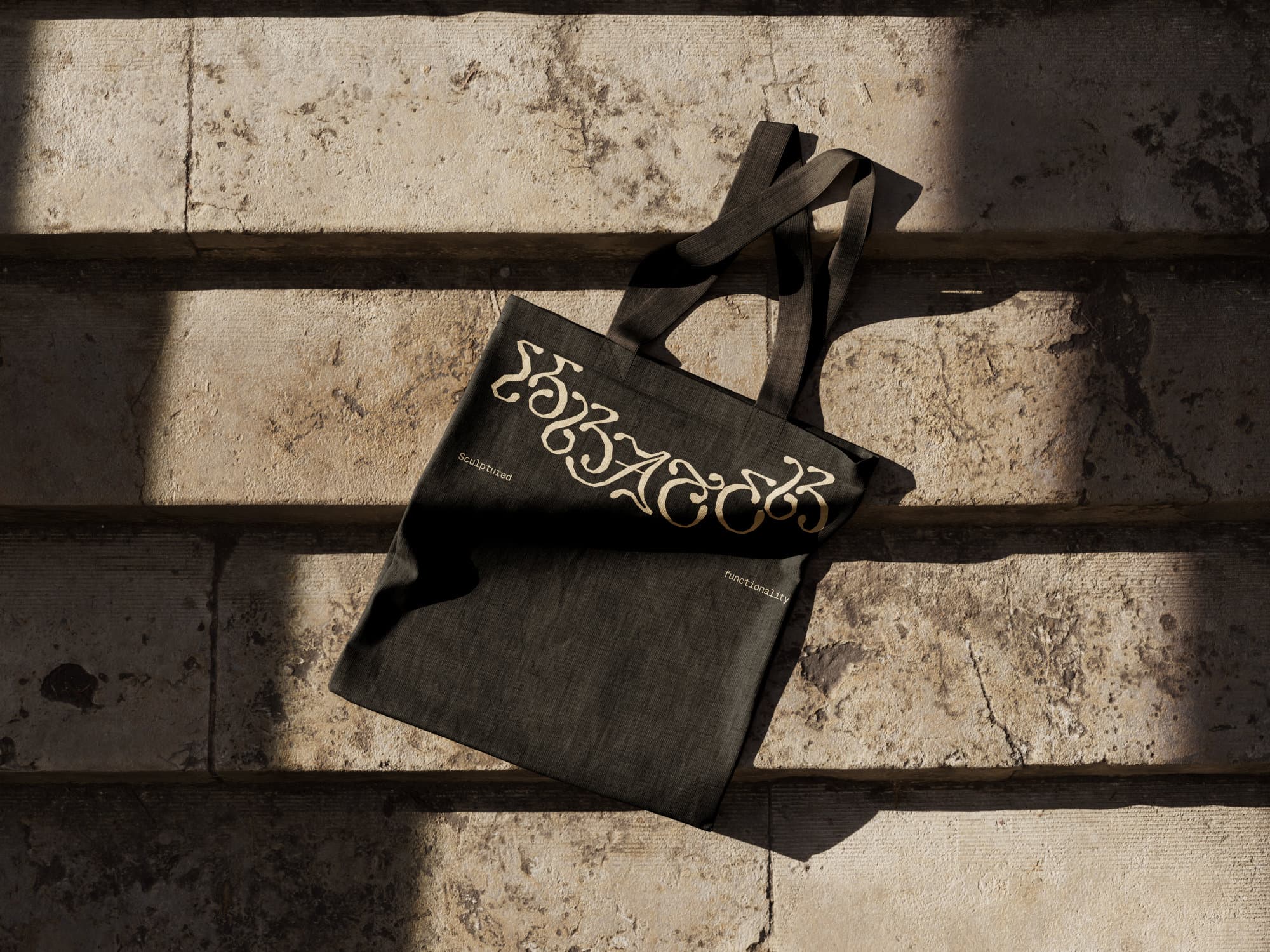 Dark canvas tote bag with the Krater.Objects wordmark in gold lettering and small text reading 'Sculptured' and 'functionality', laid across sunlit travertine steps.