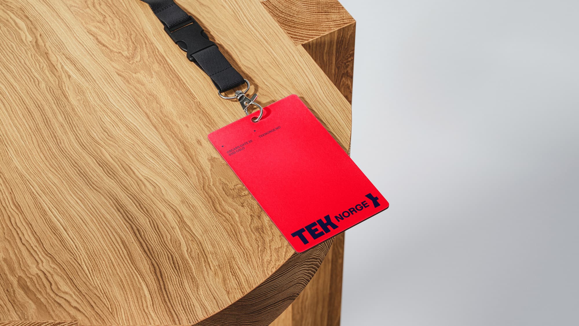 Red conference badge on a lanyard with TEK Norge's new logo, resting on an oak tabletop. Shows the brand identity applied to event materials.