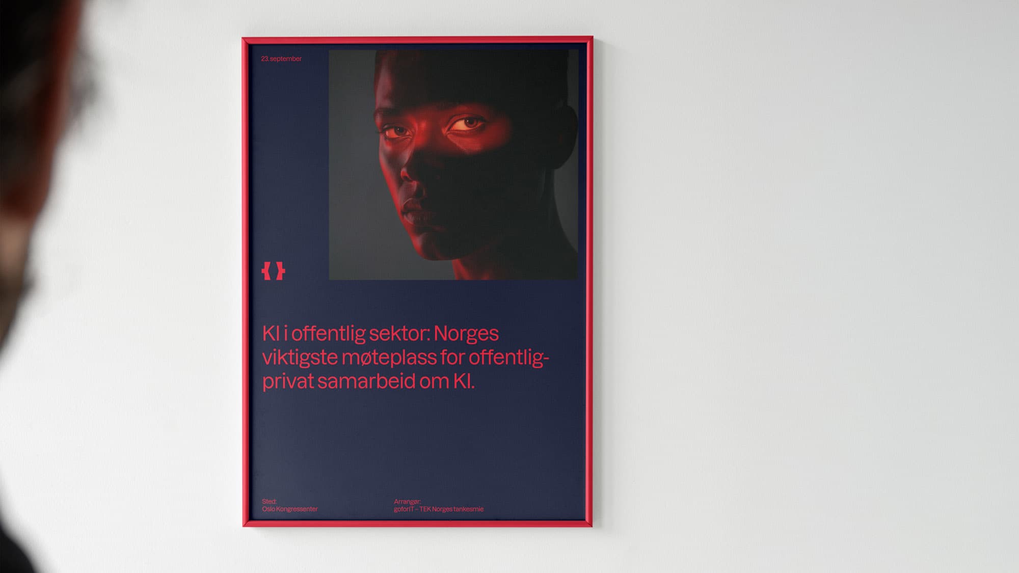 Framed TEK Norge poster on a white wall, seen over someone's shoulder in the foreground. Navy poster with a face lit in red and text: "KI i offentlig sektor: Norges viktigste møteplass for offentlig-privat samarbeid om KI."