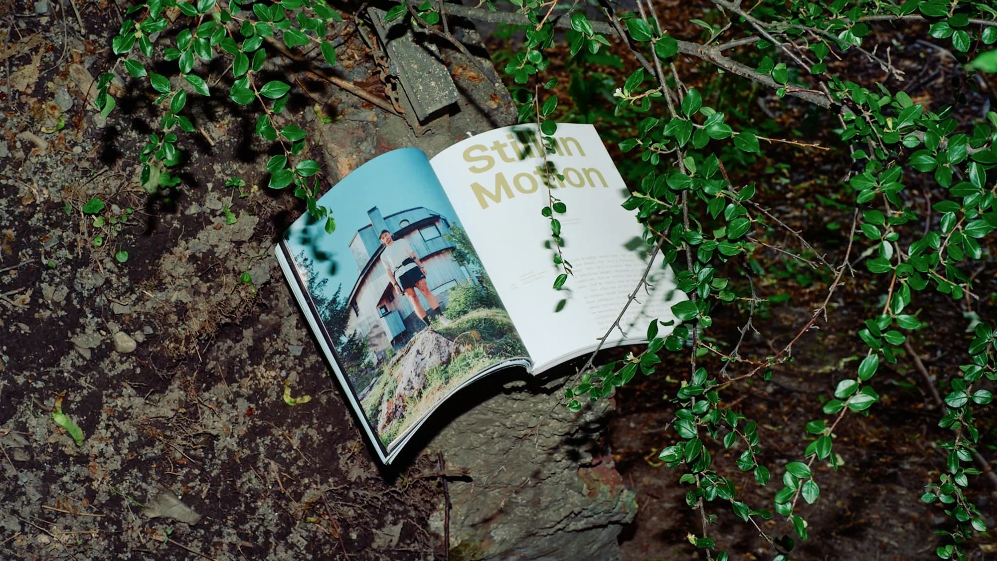 Kadens Journal open on a rock outdoors to the 'Still in Motion' spread: a runner standing on a boulder beside a blue timber cabin under open sky.