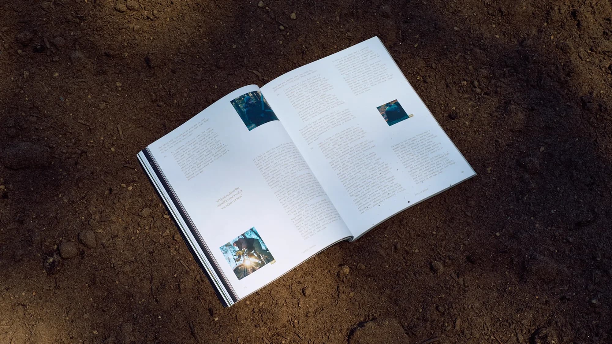 Kadens Journal photographed outdoors on dirt in raking sunlight, open to the 'Still in Motion' interview spread with forest and cycling photos and a pull quote on curiosity.