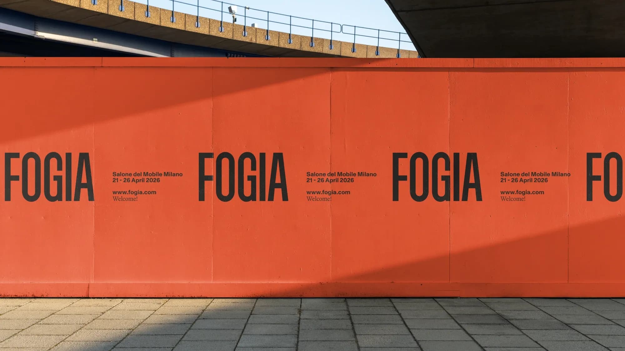 Orange construction hoarding printed with repeated FOGIA wordmarks and 'Salone del Mobile Milano, 21-26 April 2026', photographed under a road overpass.