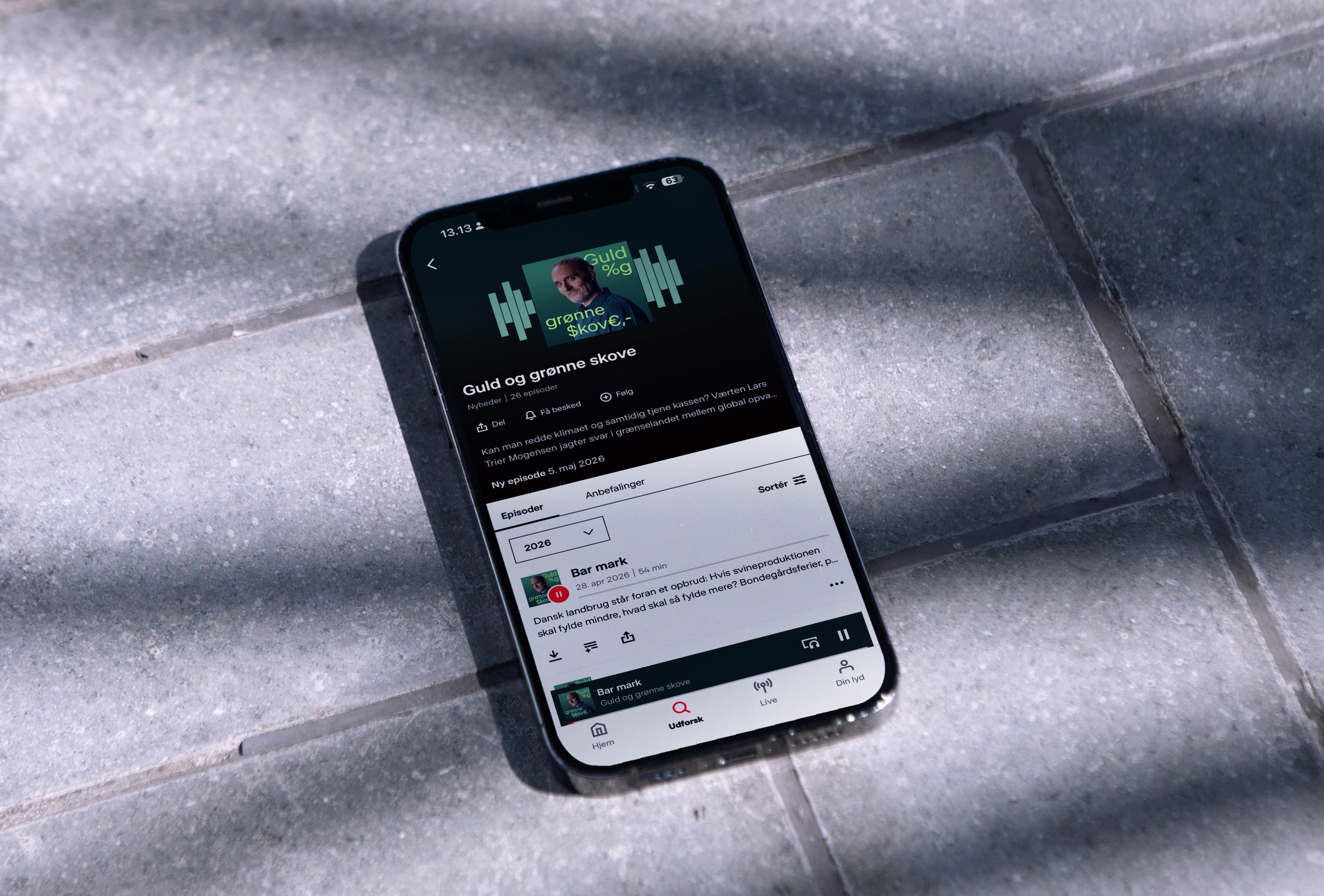 Smartphone on a stone-tiled floor showing the podcast "Guld og grønne skove" in the DR Lyd app, demonstrating the identity in digital use.
