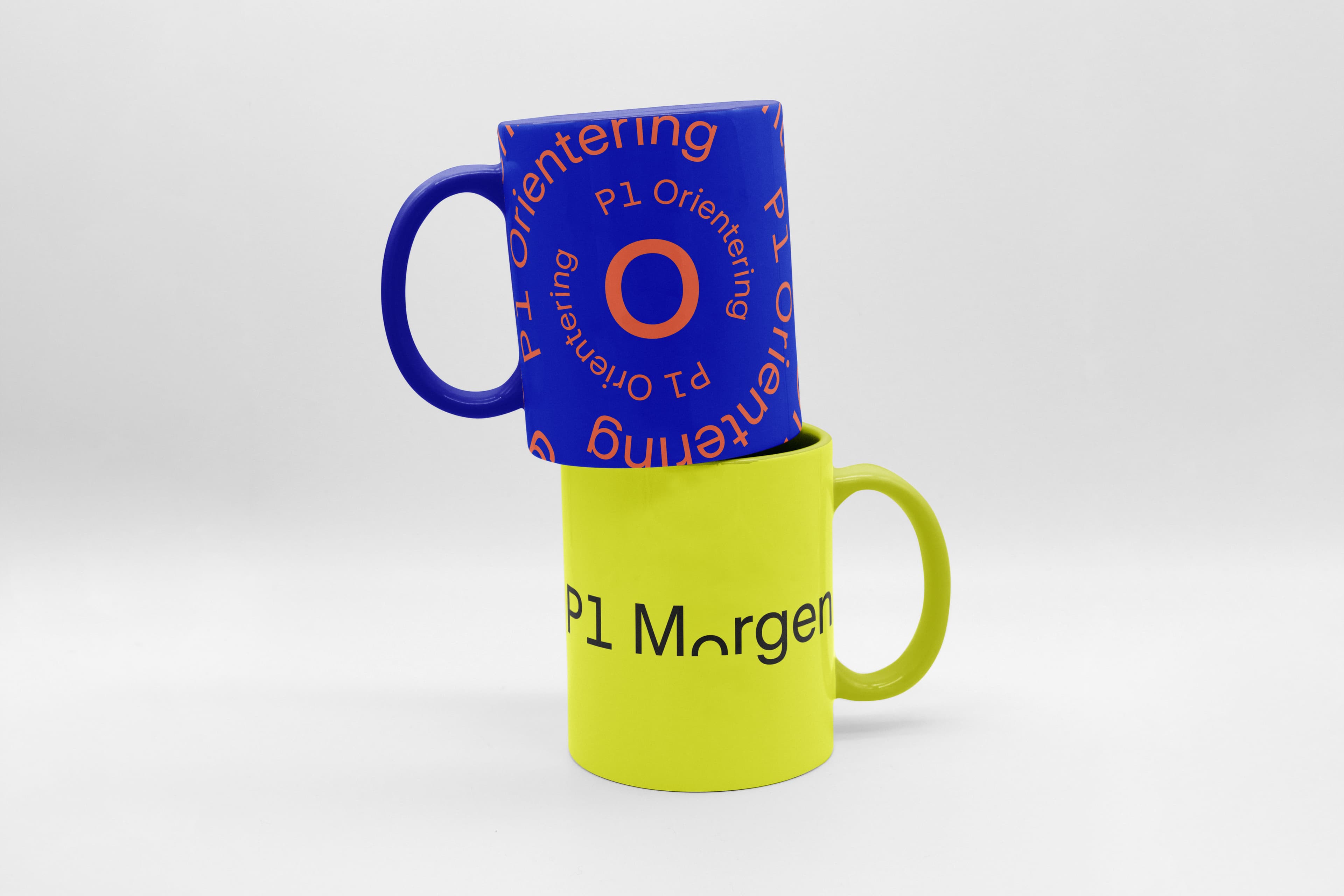 Two stacked ceramic mugs: a blue one with orange circular "P1 Orientering" text on top, and a lime-green one with black "P1 Morgen" below — brand merchandise.