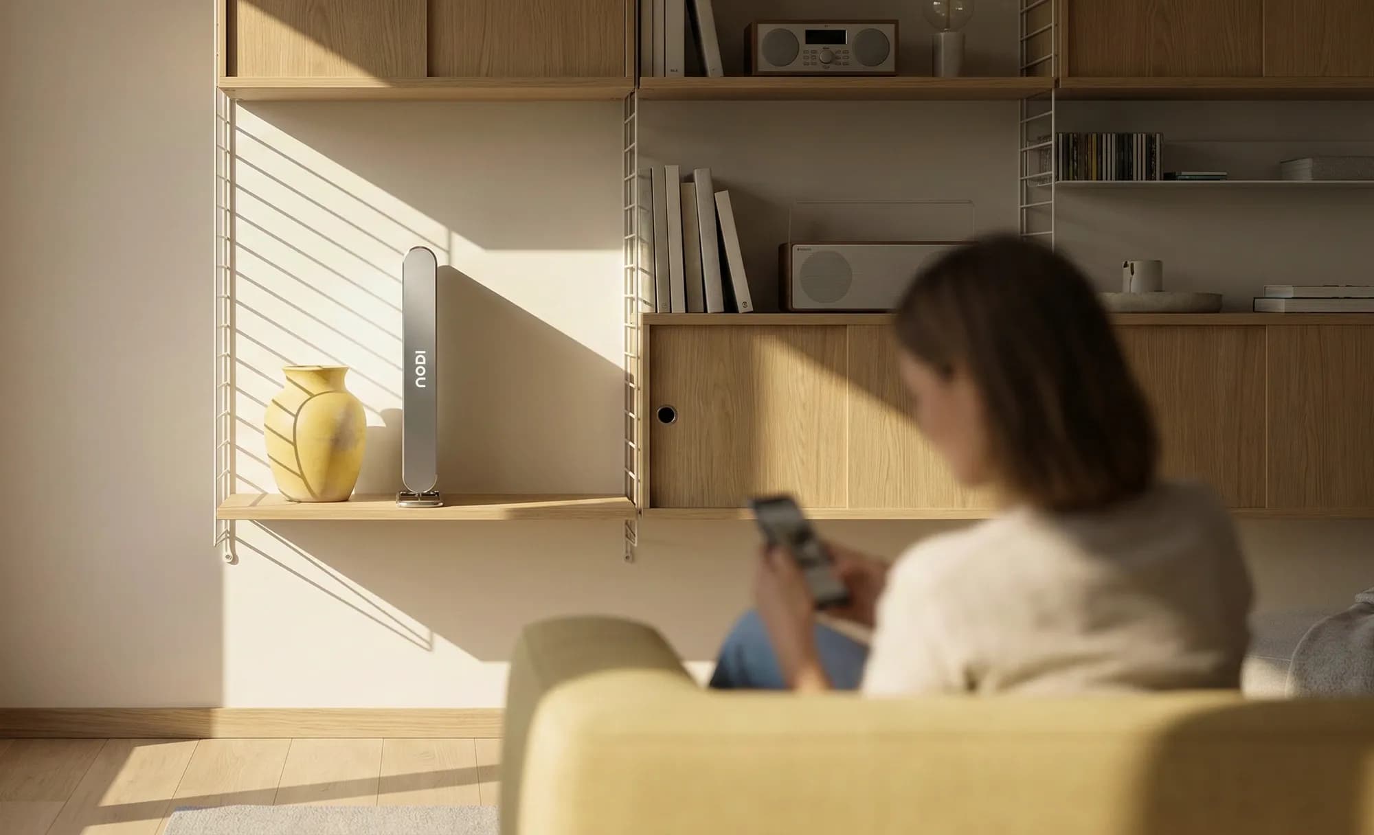 NODI battery in a bright living room: a slim metal column with the logo stands on a wooden shelf beside a yellow vase, while a blurred woman in the foreground sits on a yellow sofa using her phone. Warm sunlight and shelf shadows.