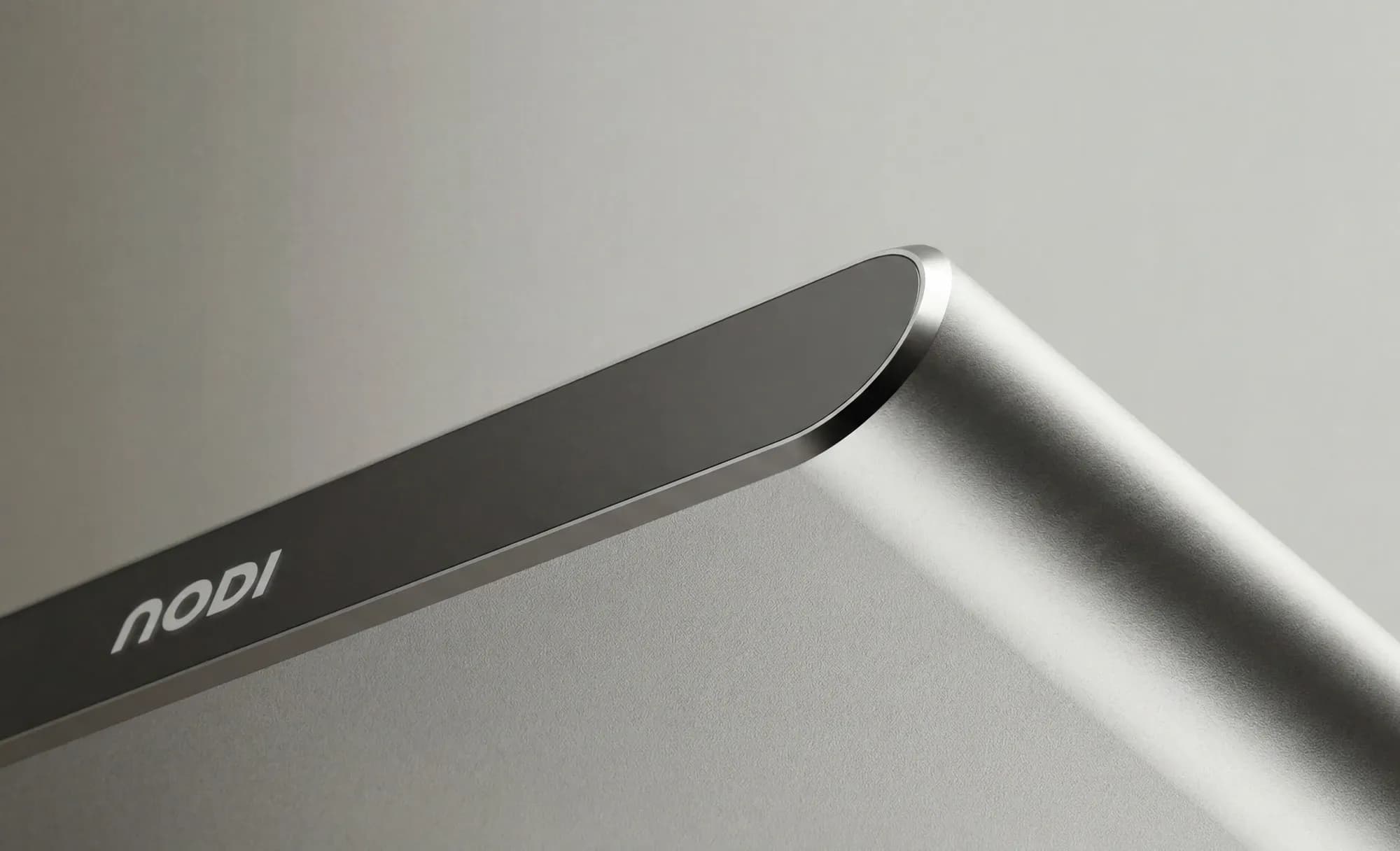Close-up of the NODI battery's rounded corner in brushed metal with a dark edge face carrying the small engraved NODI logo. A neutral light backdrop emphasises the product's clean form and material.