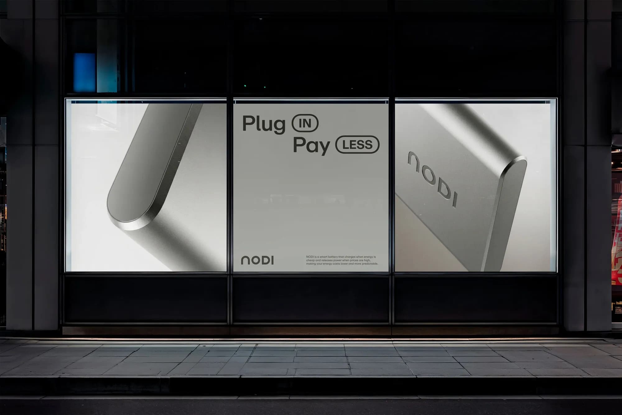Night-time shopfront window with three posters: two close-ups of the NODI battery and, in the middle, the campaign line “Plug IN, Pay LESS” with the NODI logo and a short text about charging when energy is cheap.