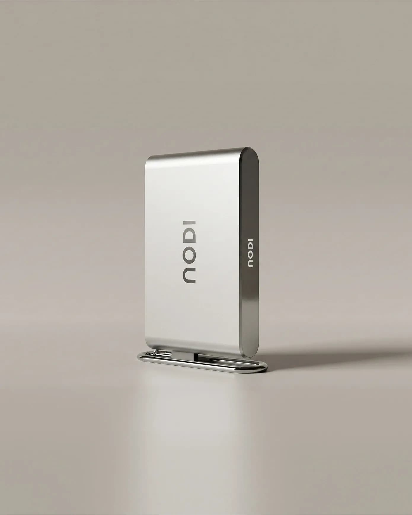 Product render of the NODI battery standing on a chrome base against a warm, neutral backdrop. The vertical logo sits on the front face and a small logo on the dark edge. Soft shadow and floor reflection.