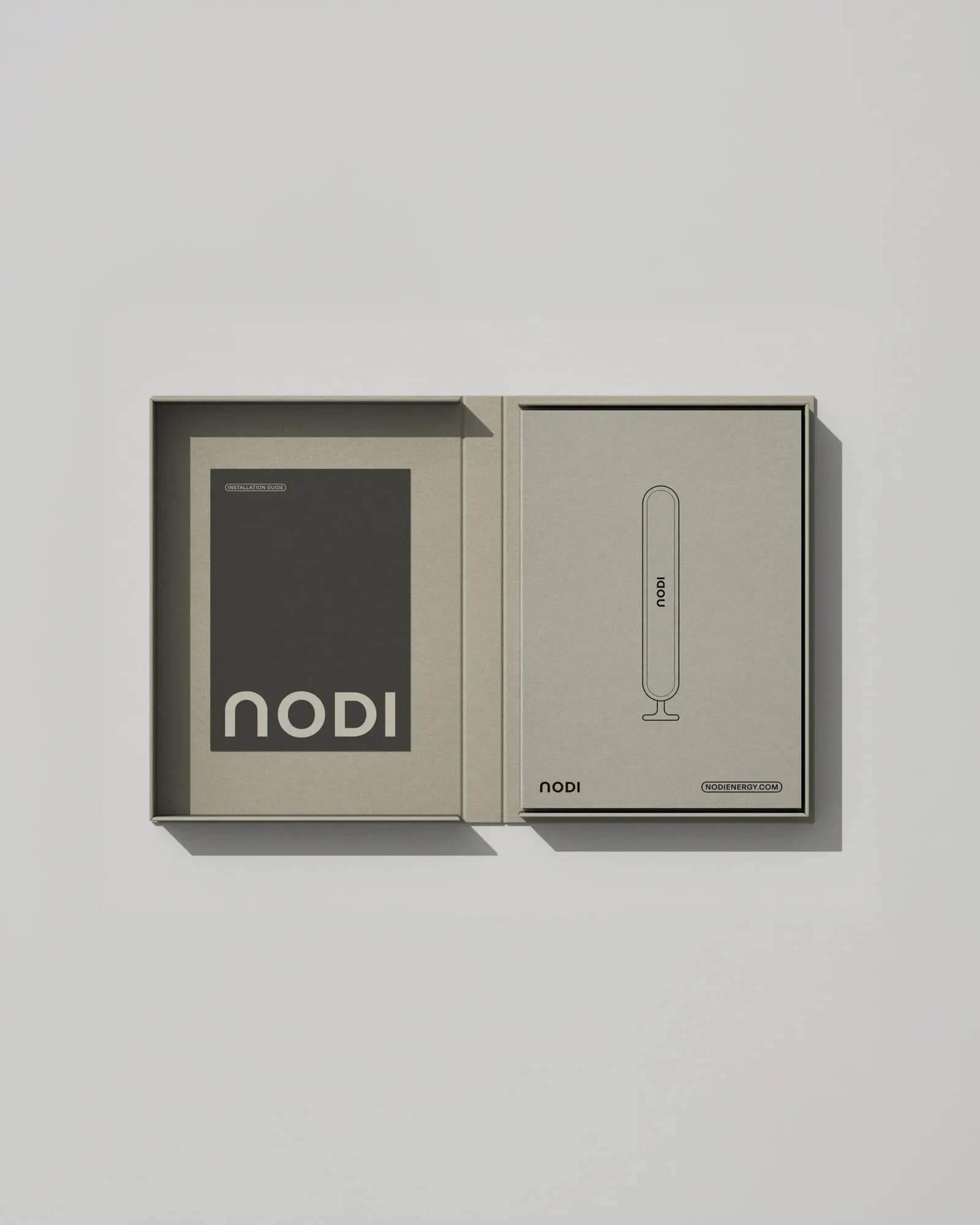 Open NODI box seen from above in vertical format: dark installation guide with the NODI logo on the left and a lid with the battery line drawing and nodienergy.com on the right. Uncoated grey-brown cardboard.