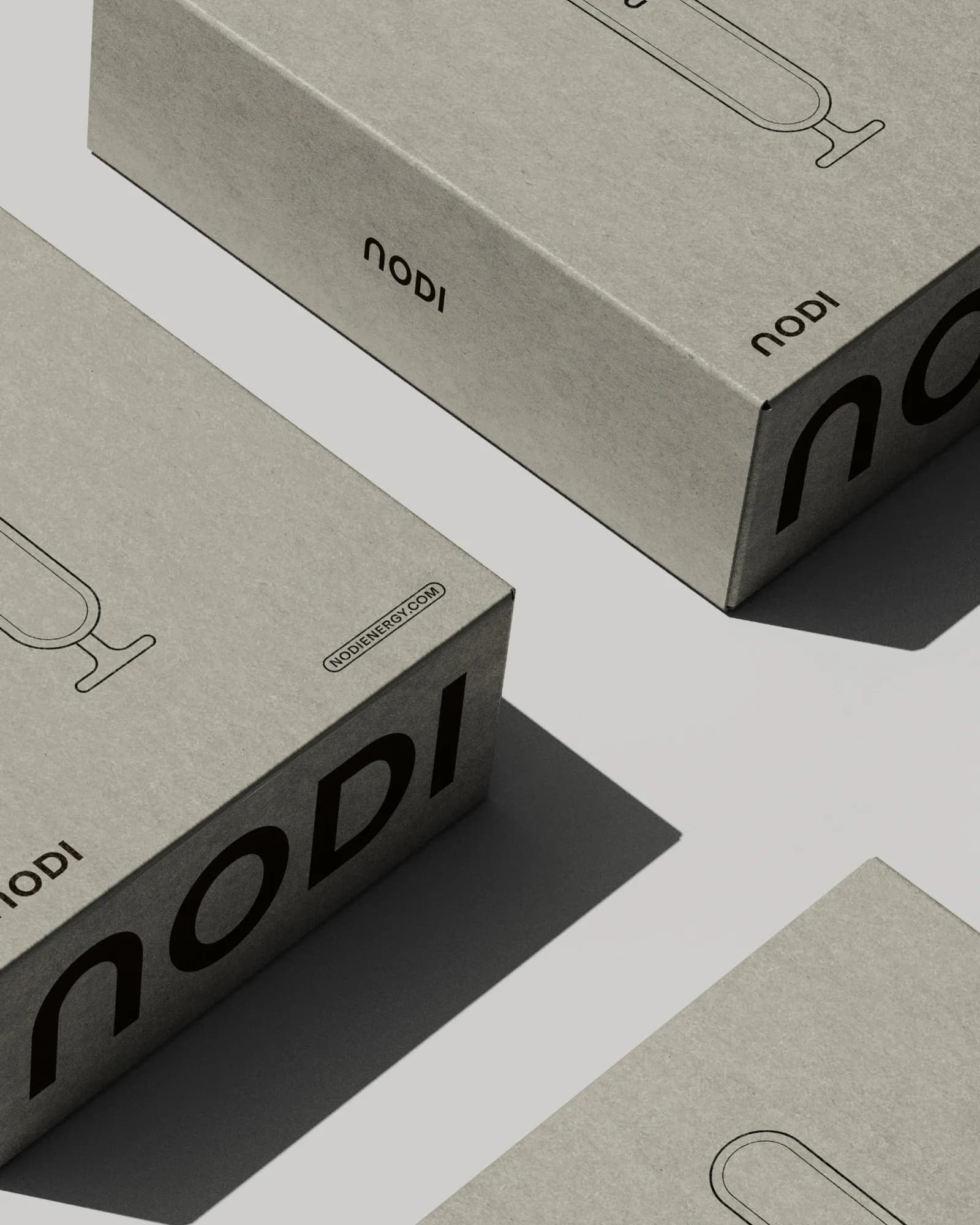 Cropped close-up of NODI boxes in grey-brown cardboard seen at an angle: oversized black logo along the edge, small logo, URL label and the battery line drawing. Hard shadows on a light grey surface.