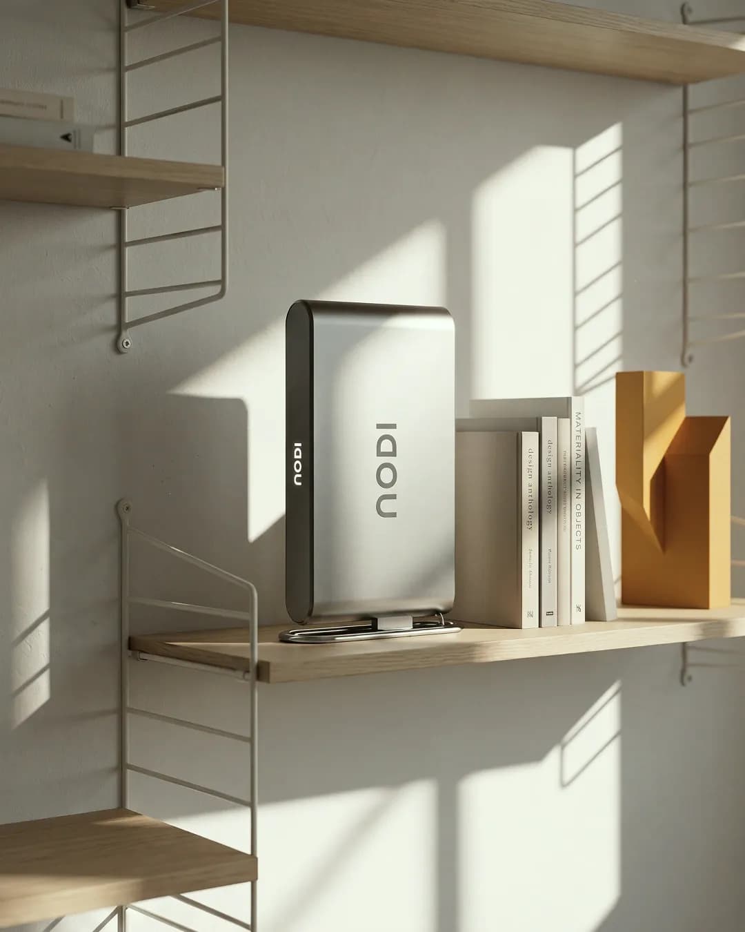NODI battery on a wooden shelf in a modular shelving system, beside stacked design books and a yellow paper sculpture. Sharp sunlight casts patterns from the shelf rails across the light wall. Vertical format.