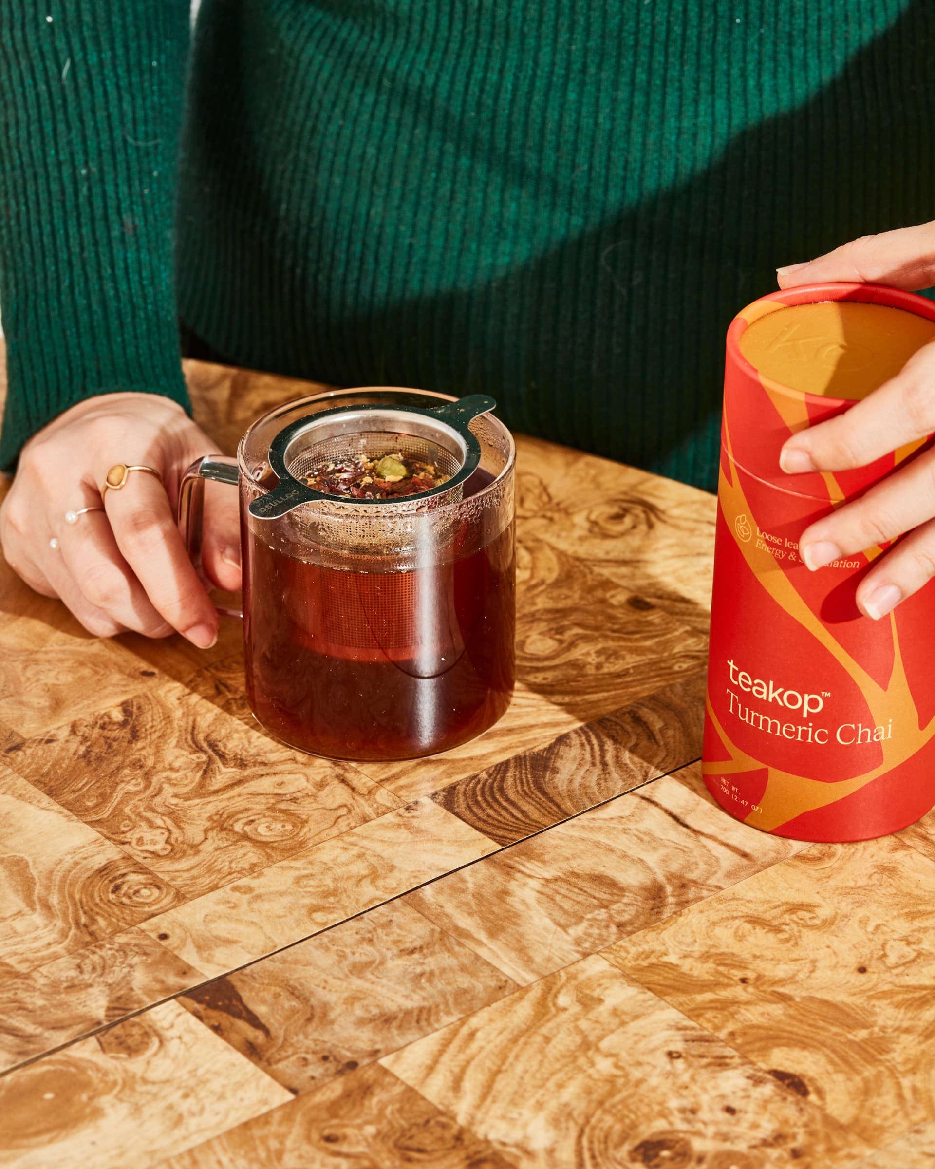 A person wearing a green sweater and holding a red and yellow pack of tea from teakop.