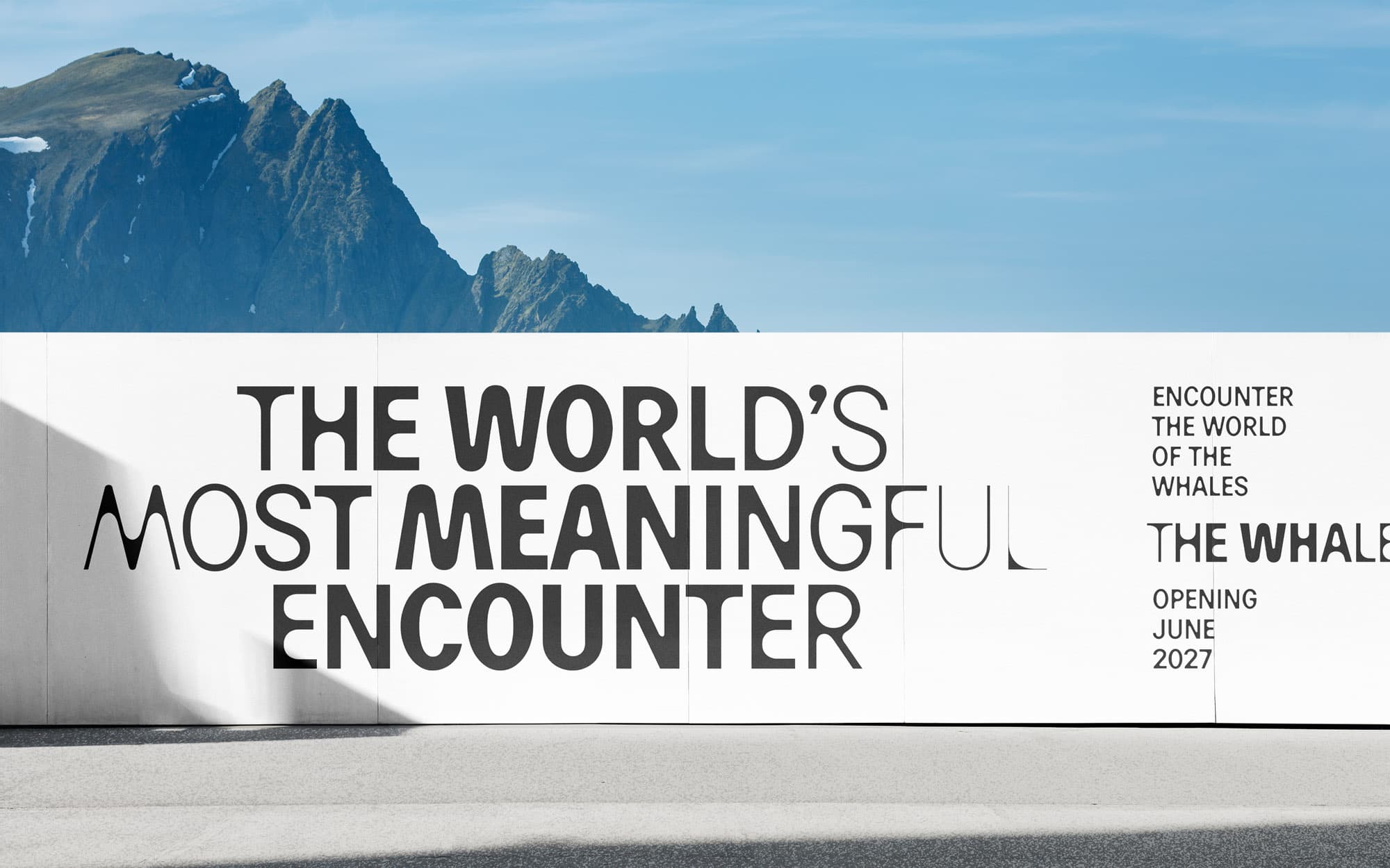 Large wall mockup: white advertising hoarding with "The World's Most Meaningful Encounter – The Whale, Opening June 2027" in black type; Norwegian mountain peaks rising above the wall.
