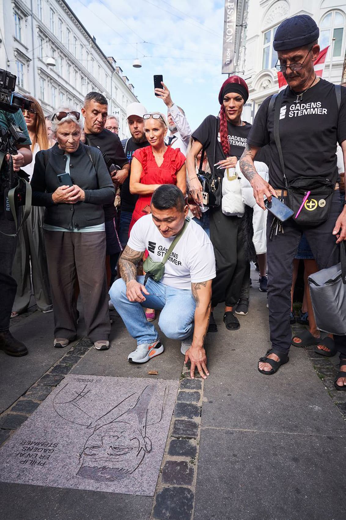 Man in a Gadens Stemmer t-shirt crouches to touch his engraved sidewalk stone, surrounded by onlookers and press at the unveiling ceremony