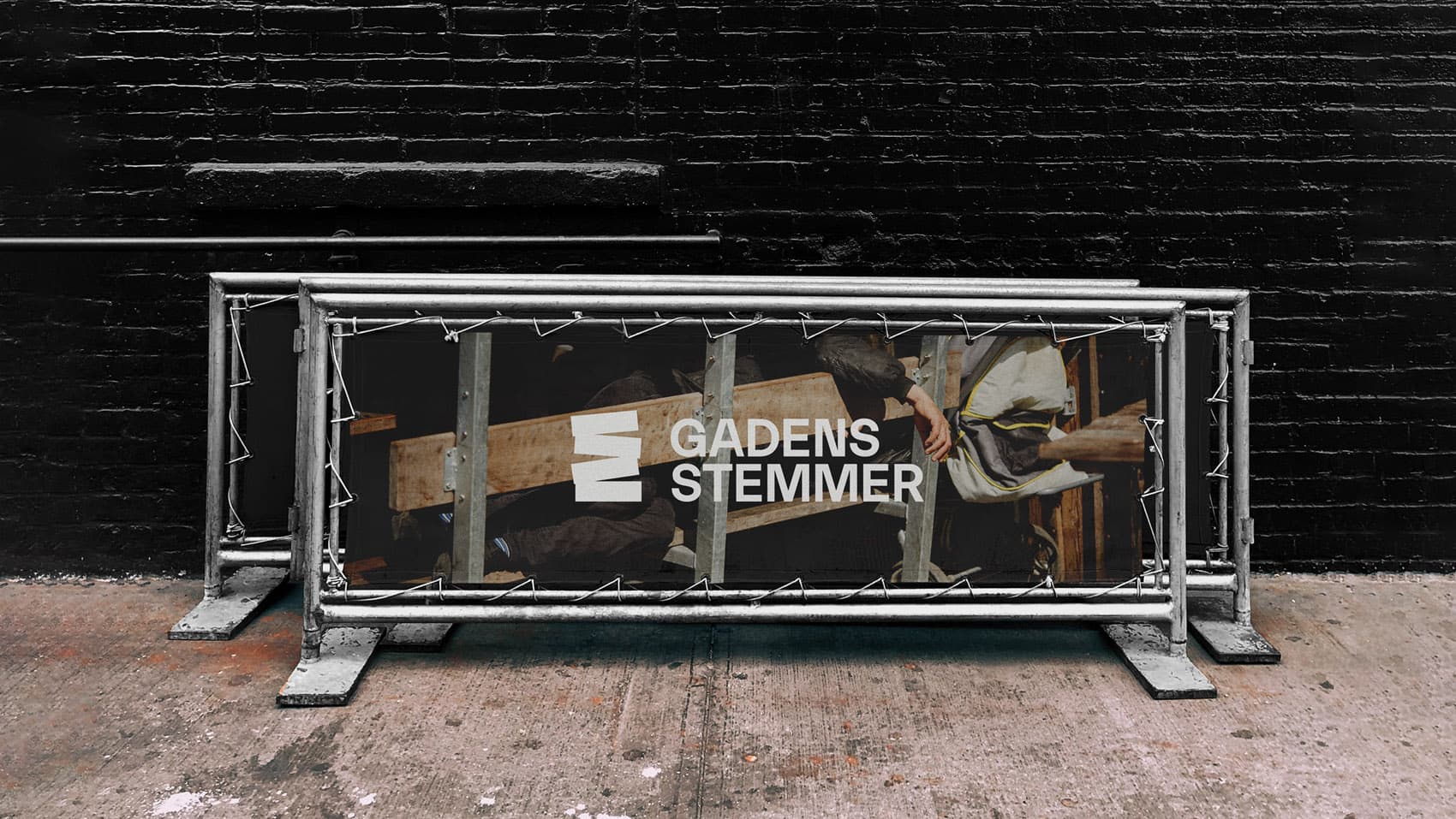Metal street barrier featuring the Gadens Stemmer logo and classical painting imagery, placed against a dark brick wall