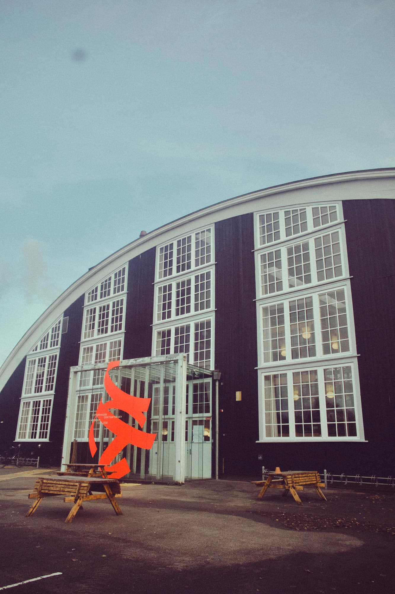 Black-painted hangar building with tall white window frames and CCCC's orange logo placed by the entrance — the school's main facade.