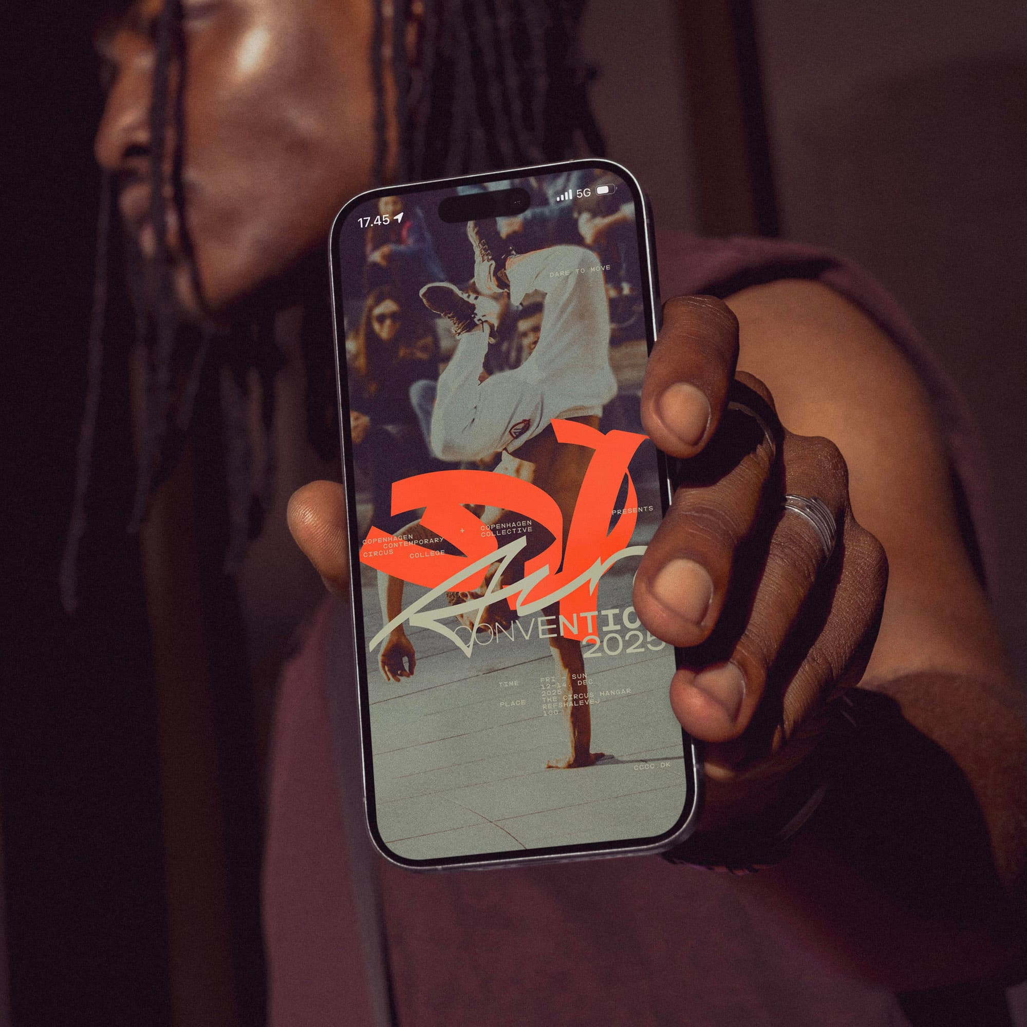 iPhone in hand shows CCCC's poster for 'Cir Convention 2025' with orange custom typography over a breakdance photo; the owner is softly out of focus behind.