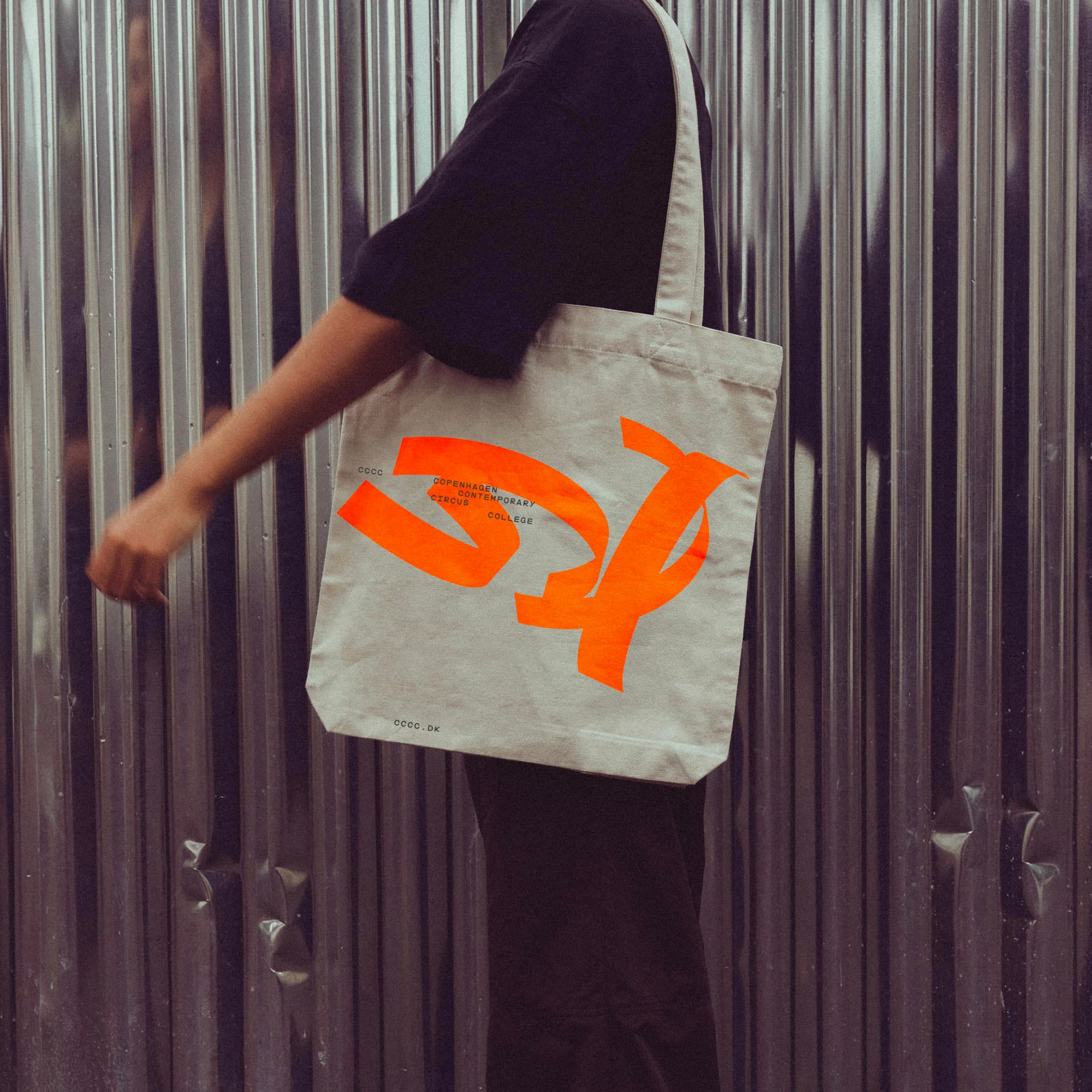 Person carries a CCCC canvas tote bag in raw white with large orange logo, photographed against a corrugated metal wall.