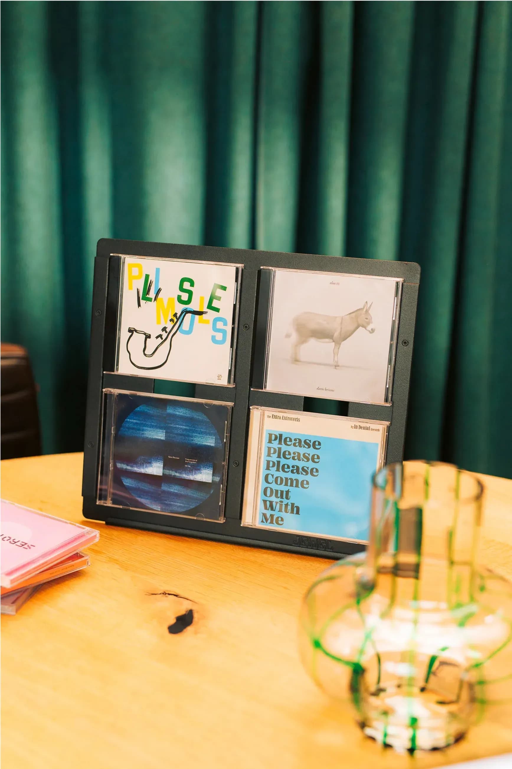 The Jewel CD rack placed on a table surrounded by glass in front of a green window drape.
